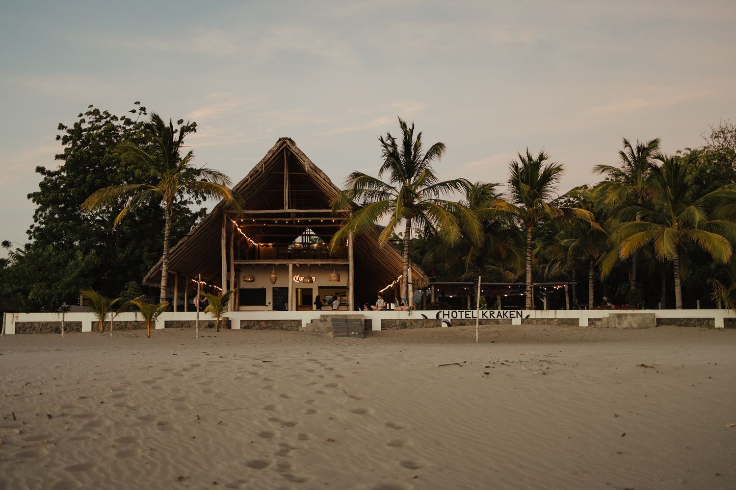 Beachfront thatched-roof restaurant surrounded by palm trees on a white sandy tropical beach at dusk.