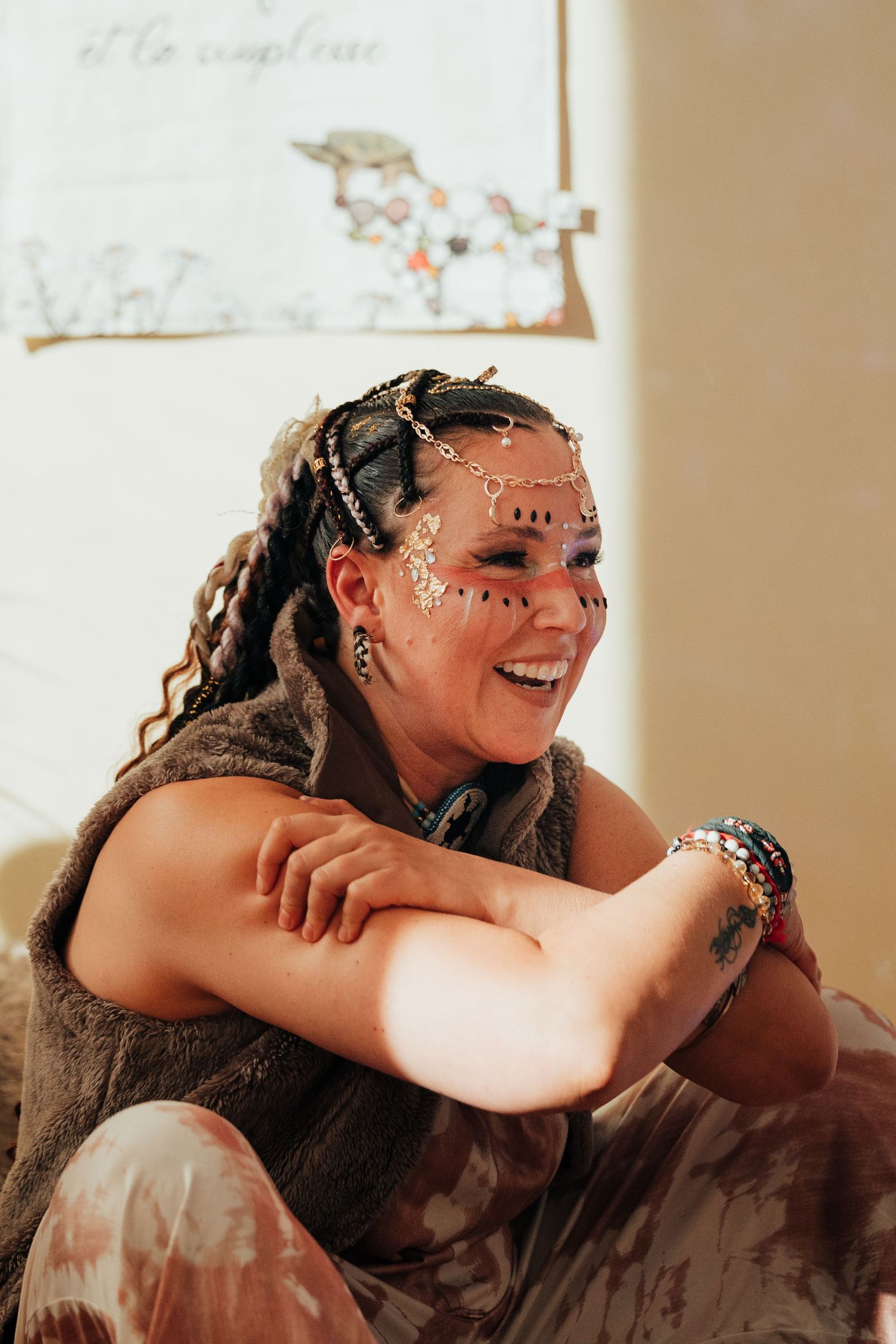 Smiling woman with braided hair, floral face gems, colorful bracelets, and fur vest sits cross-armed in warm sunlight.