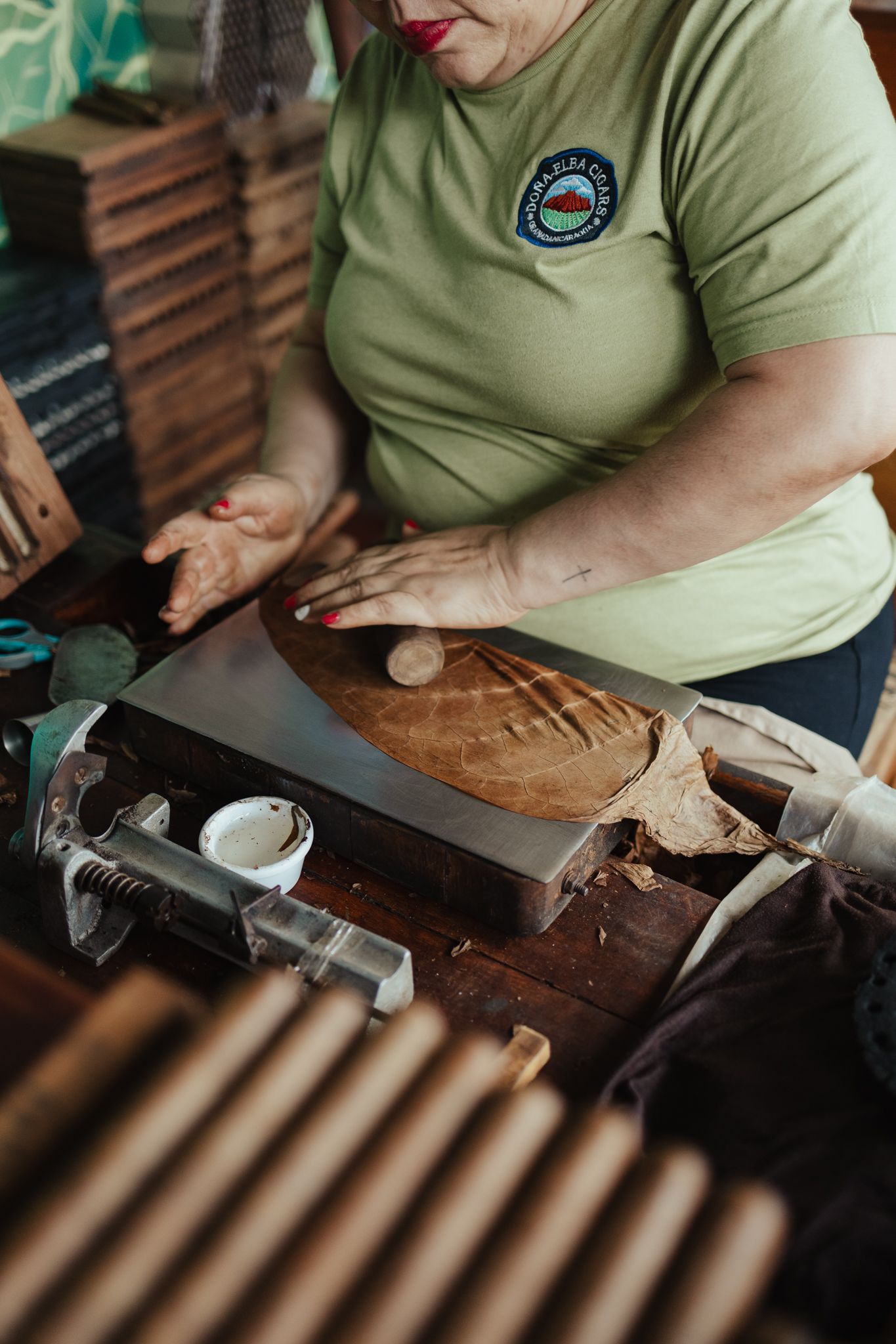 A skilled woman in a green t-shirt hand-rolling cigars at a wooden workbench in a traditional cigar factory.