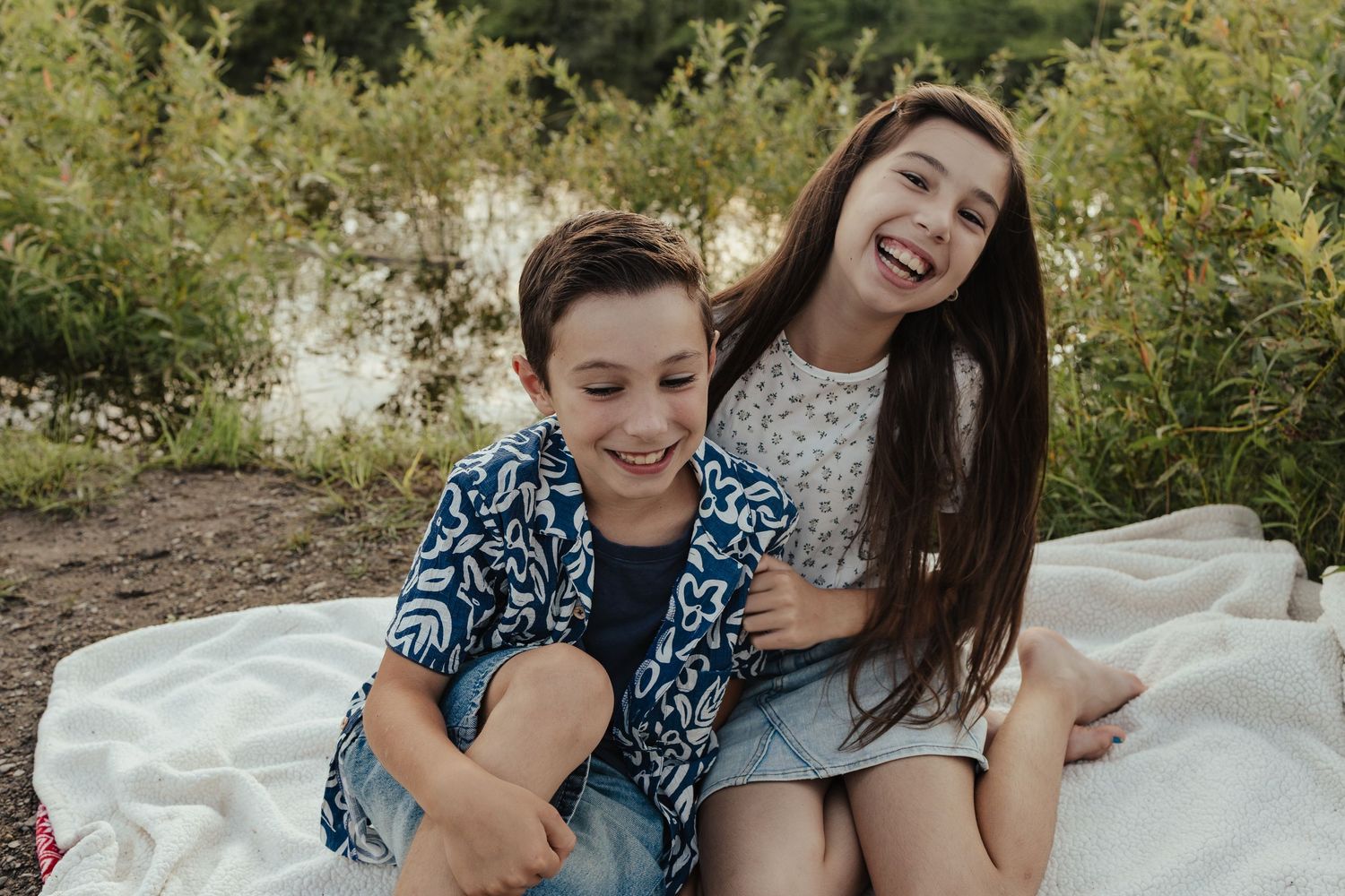 Two happy children sitting on a blanket outdoors near a lake, smiling and laughing together in casual summer clothing.