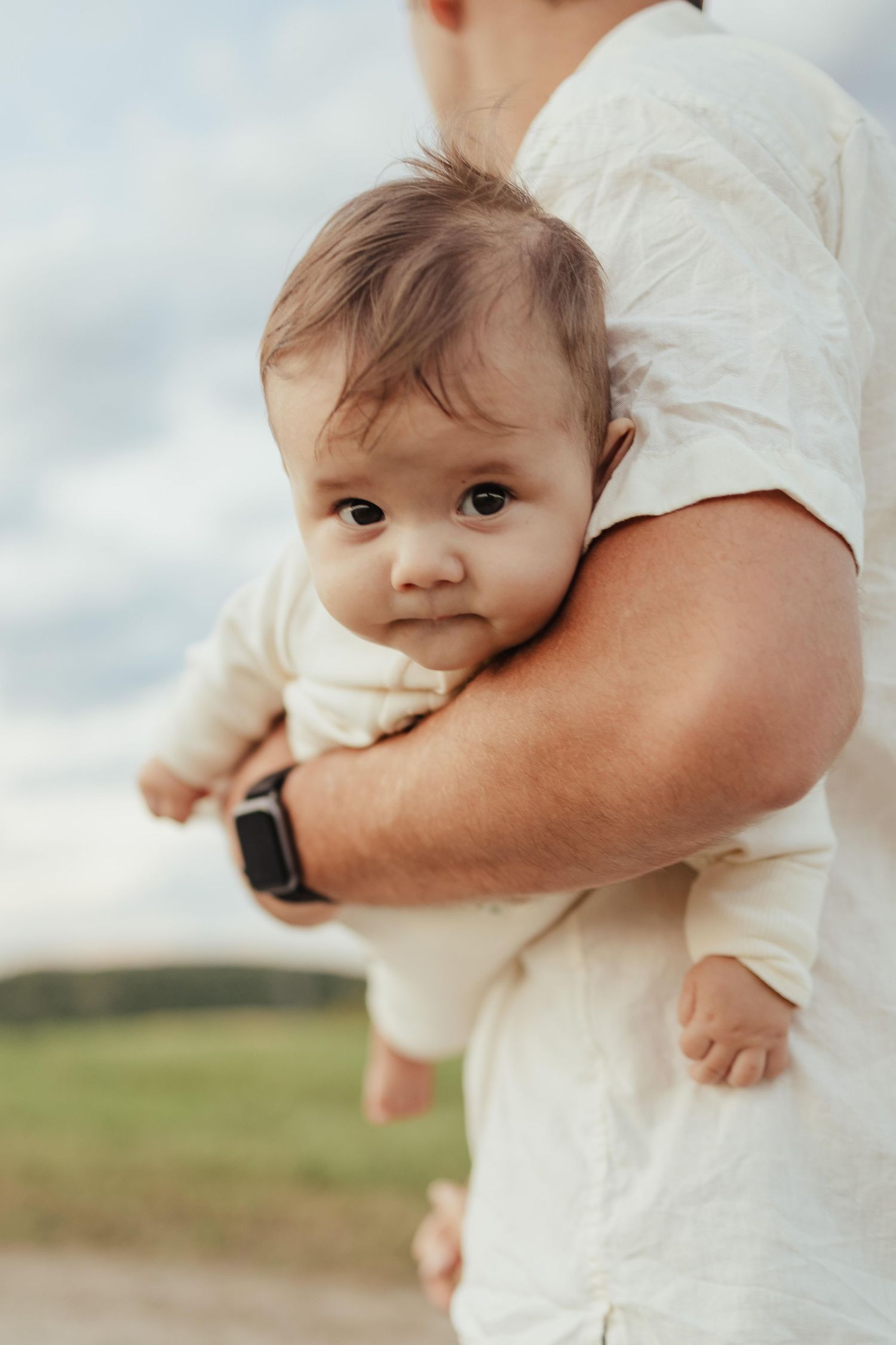Father holding baby outdoors in white linen shirt with smartwatch, infant gazing at camera against cloudy sky.