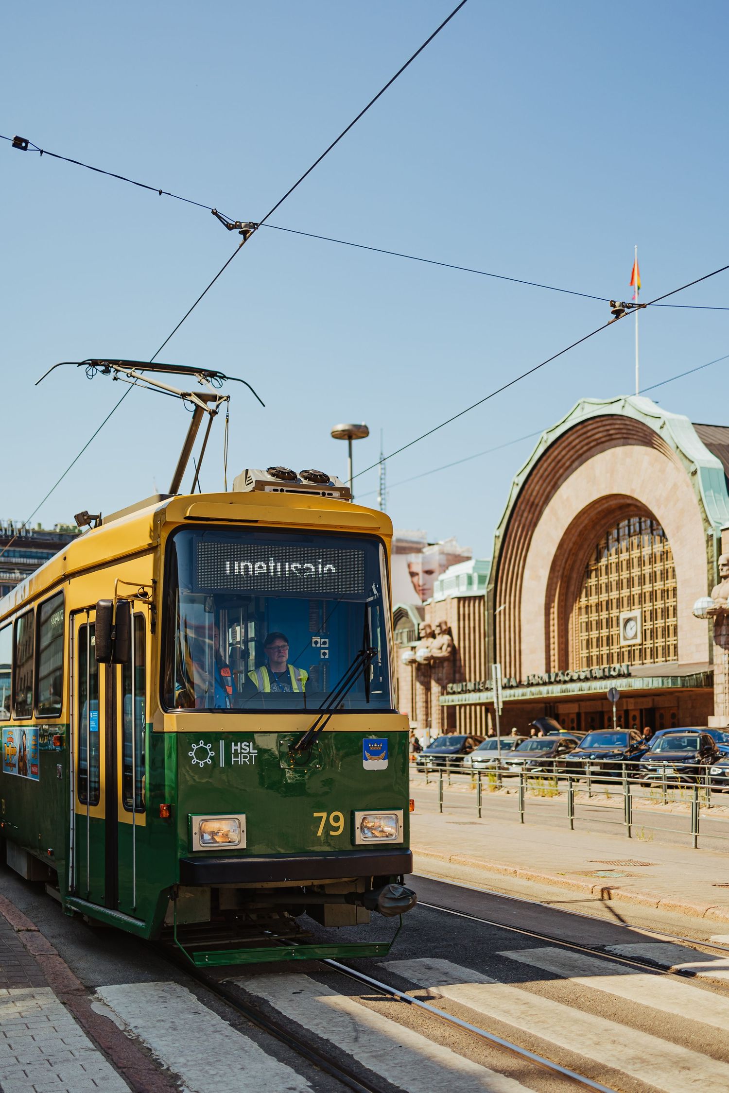 A green and yellow tram number 79 travels past Helsinki Central Railway Station on a clear sunny day.