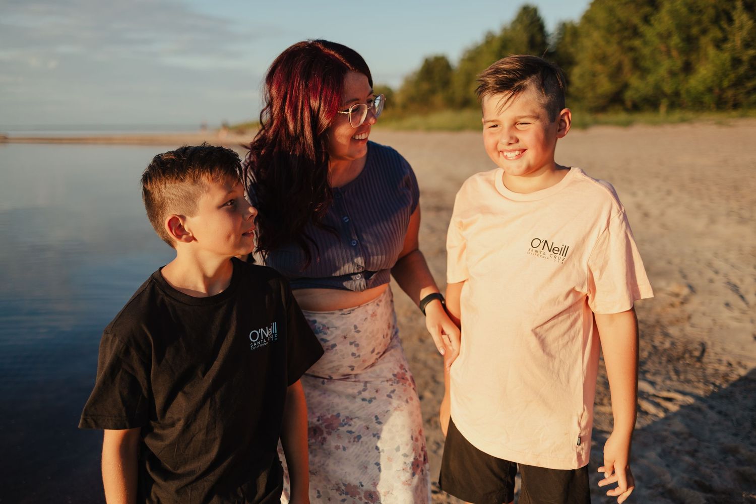 A woman and two boys wearing O'Neill shirts smile together on a sunny beach shoreline.
