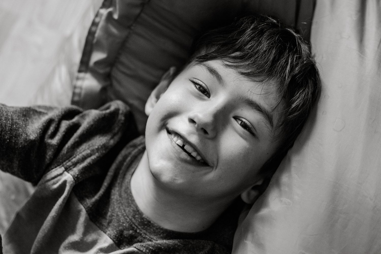 Black and white portrait of a smiling young boy lying down, looking up at the camera with a joyful expression.