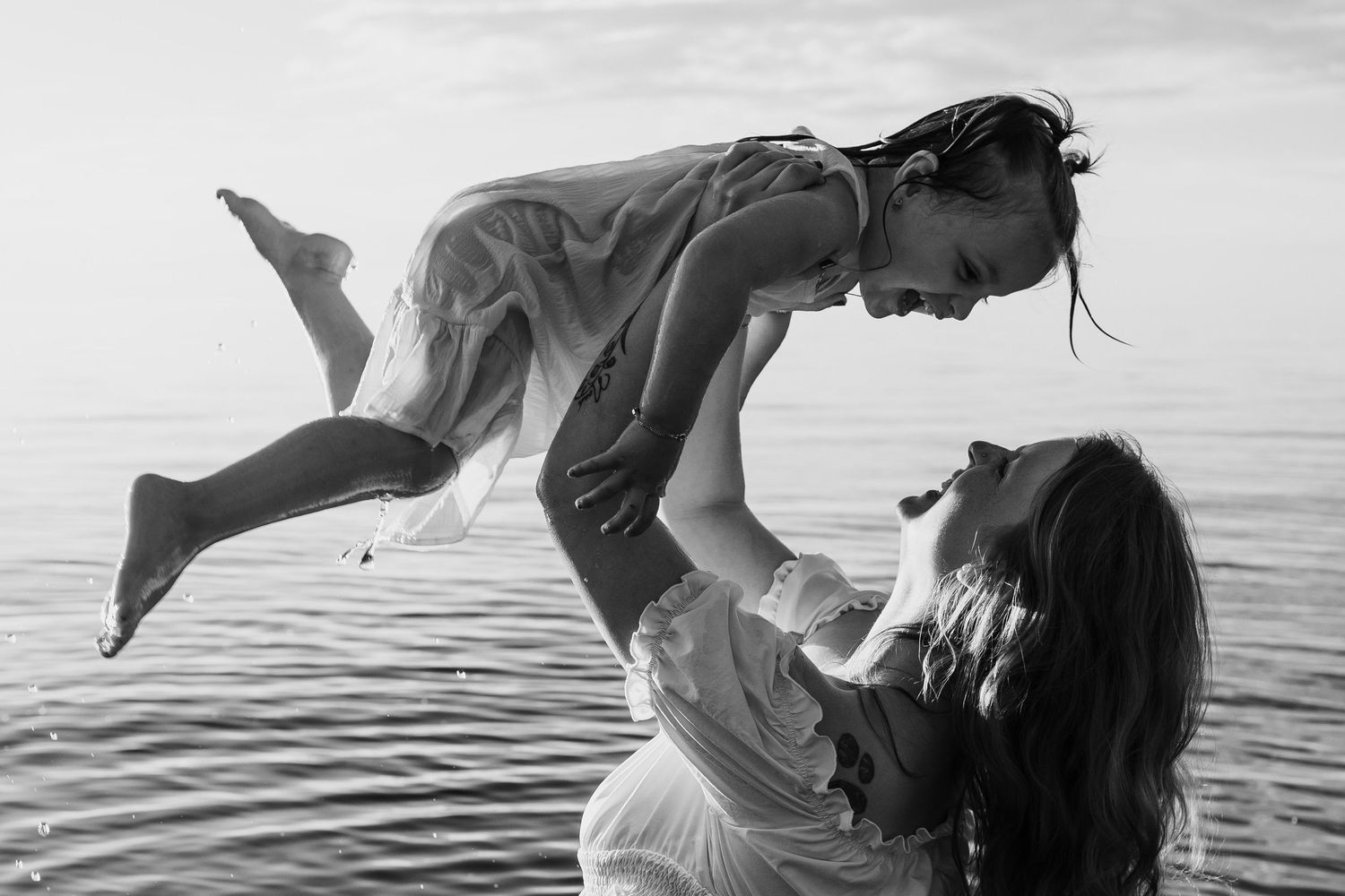 Black and white photo of mother lifting laughing child above water at beach in joyful moment.