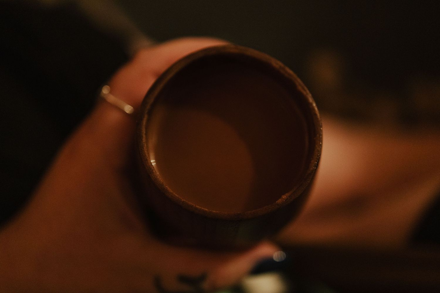 A hand holding a dark ceramic mug filled with coffee or hot chocolate in a dimly lit setting.