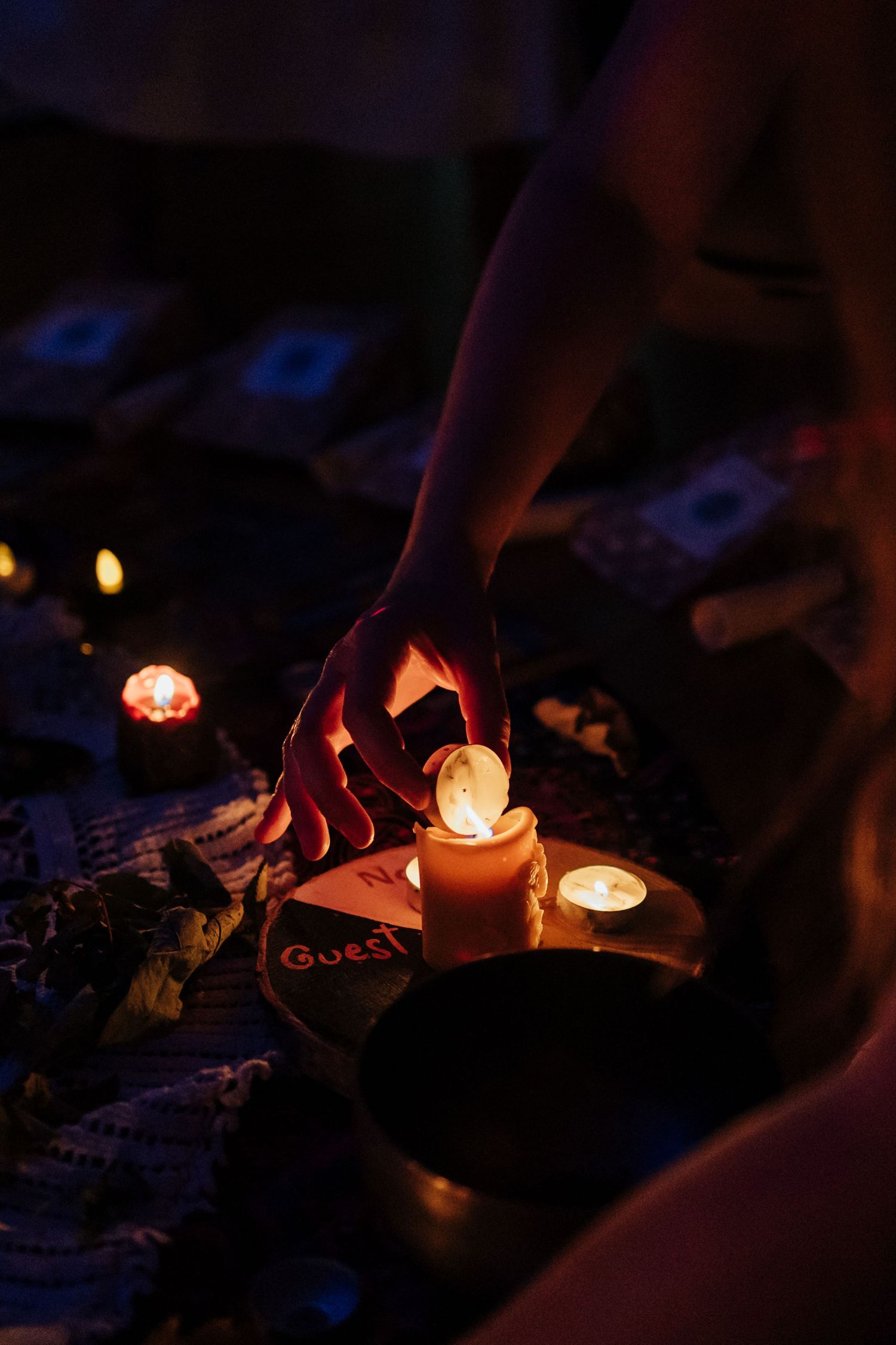 Hand touching a lit candle surrounded by tarot cards, crystals, and small candles in a dark spiritual setting.