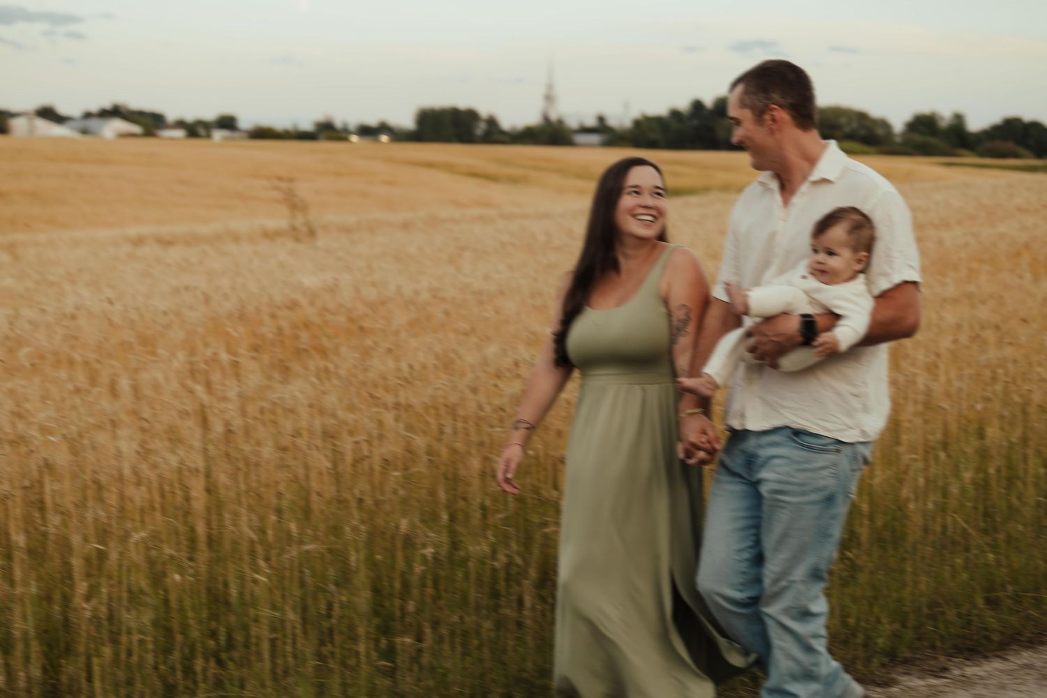 Happy family with baby walking through golden wheat field at sunset, smiling couple enjoying outdoor nature stroll.