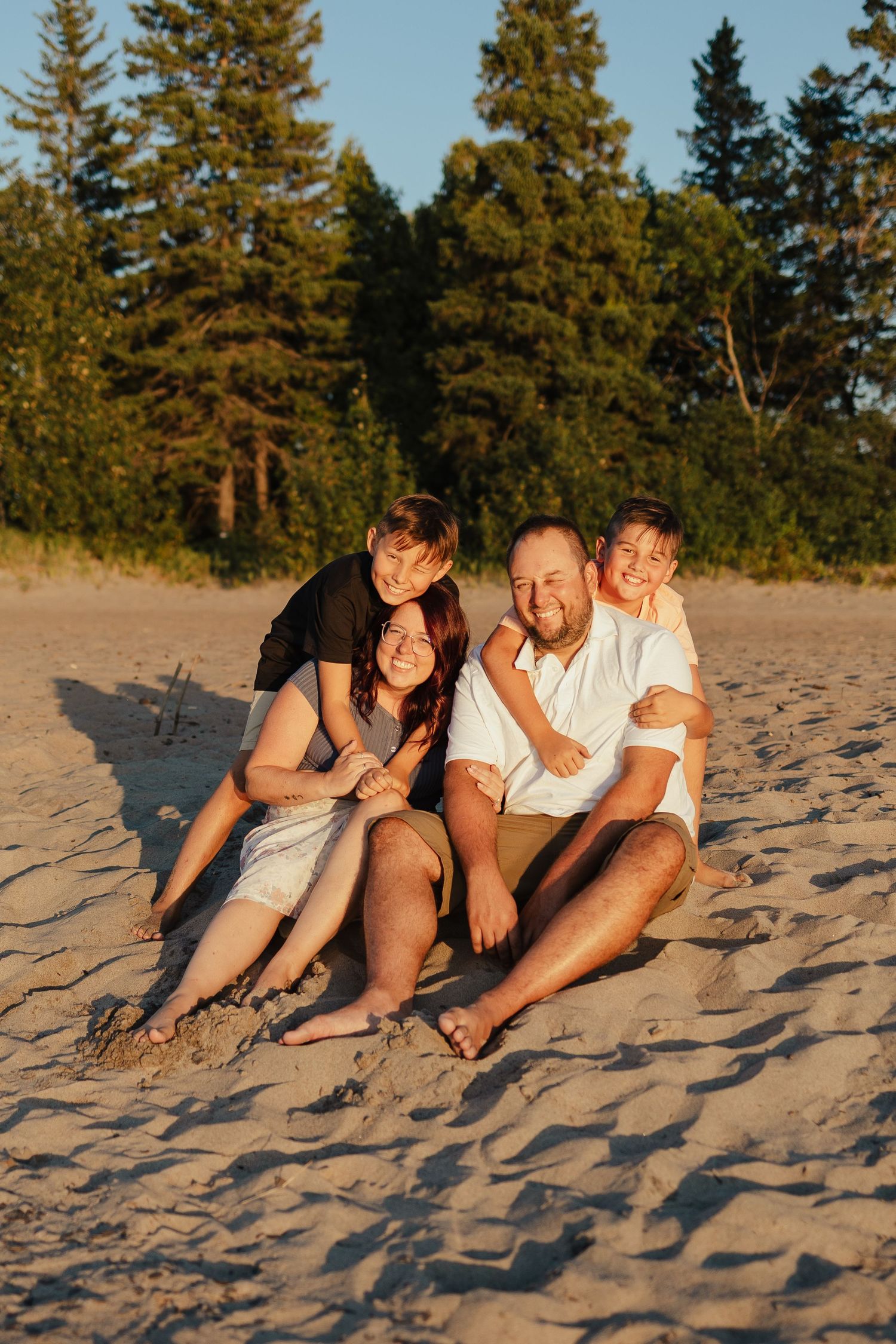 Family of four smiling and sitting together on sandy beach at golden hour with pine trees in background.