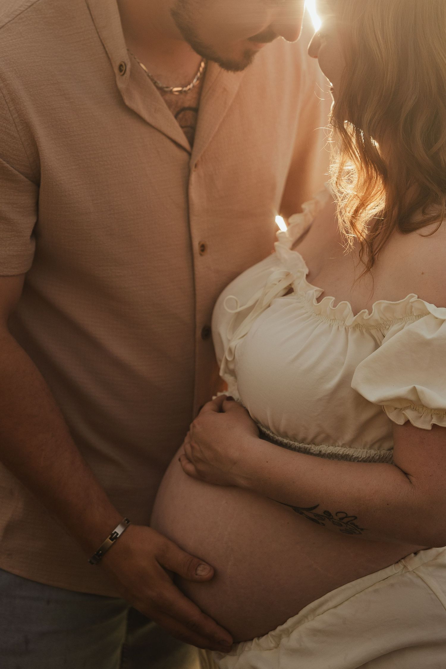 Couple sharing an intimate moment in golden sunlight, cradling a pregnant belly outdoors.