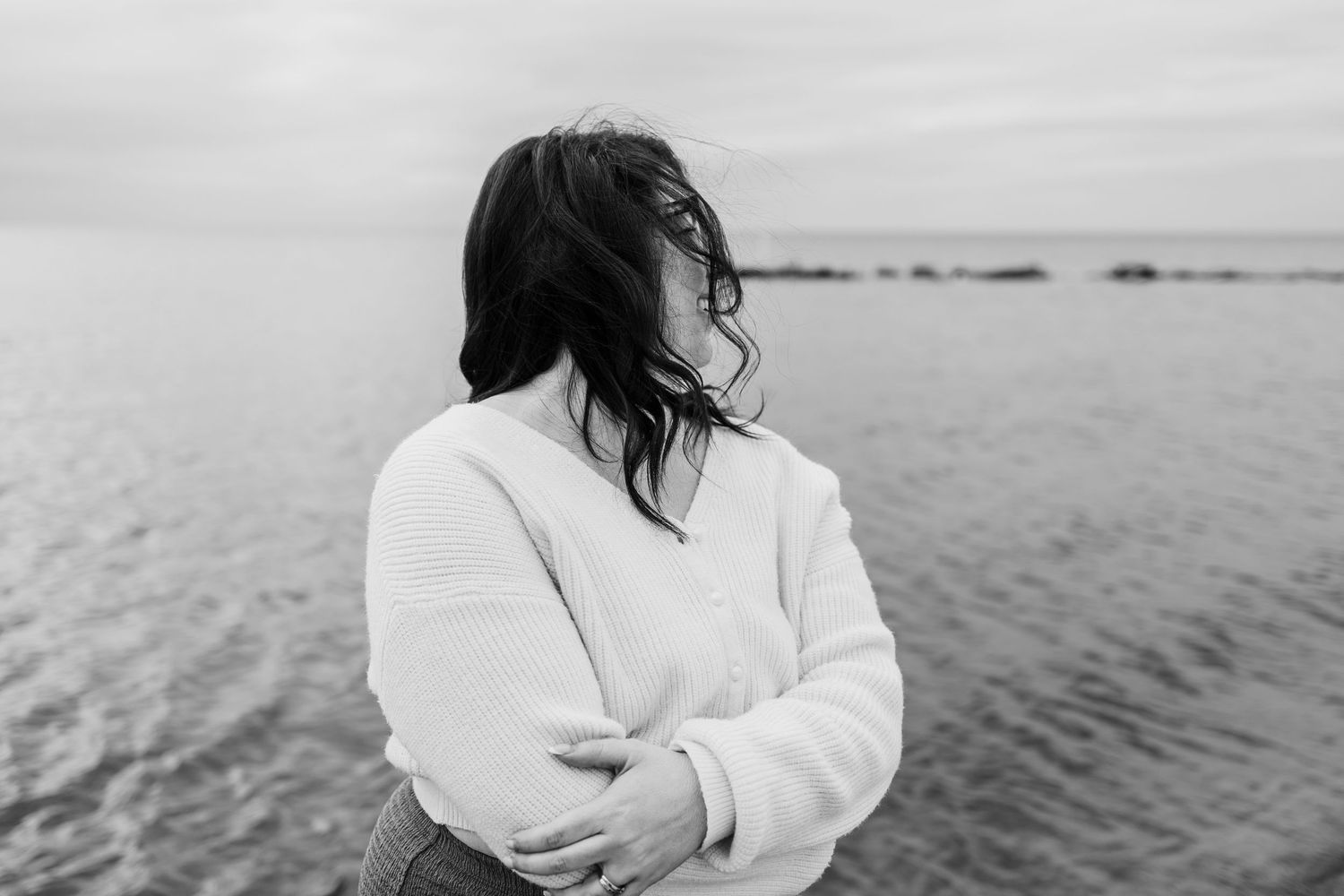 Black and white photo of a woman in a white sweater standing by the ocean with wind blowing her dark hair.