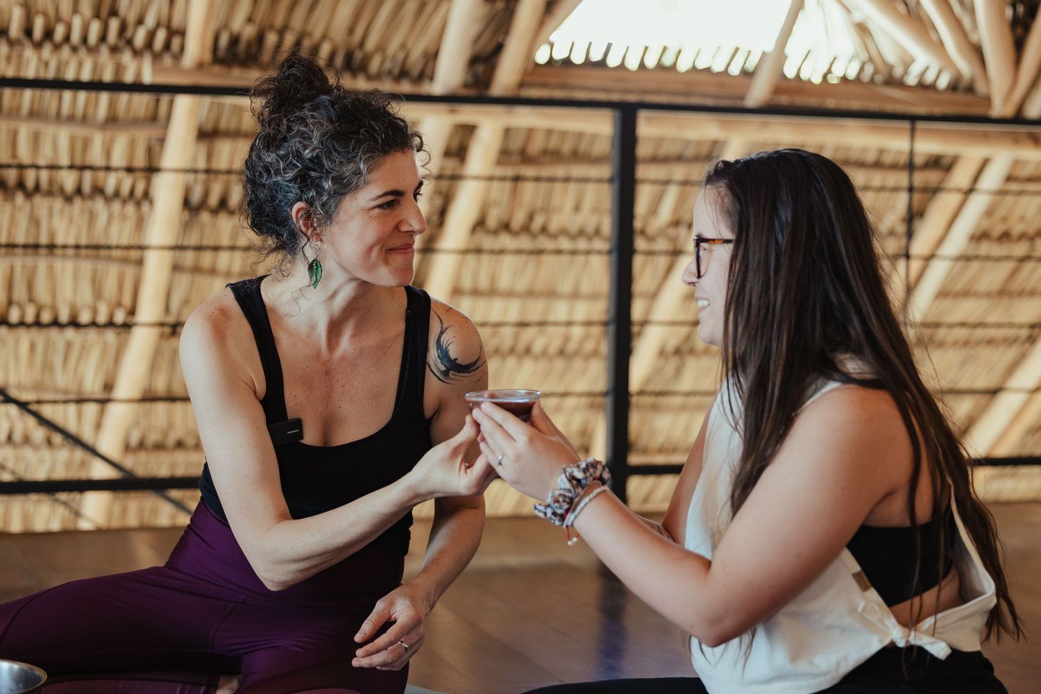 Two women sharing a mindful conversation in a serene thatched-roof yoga studio setting.