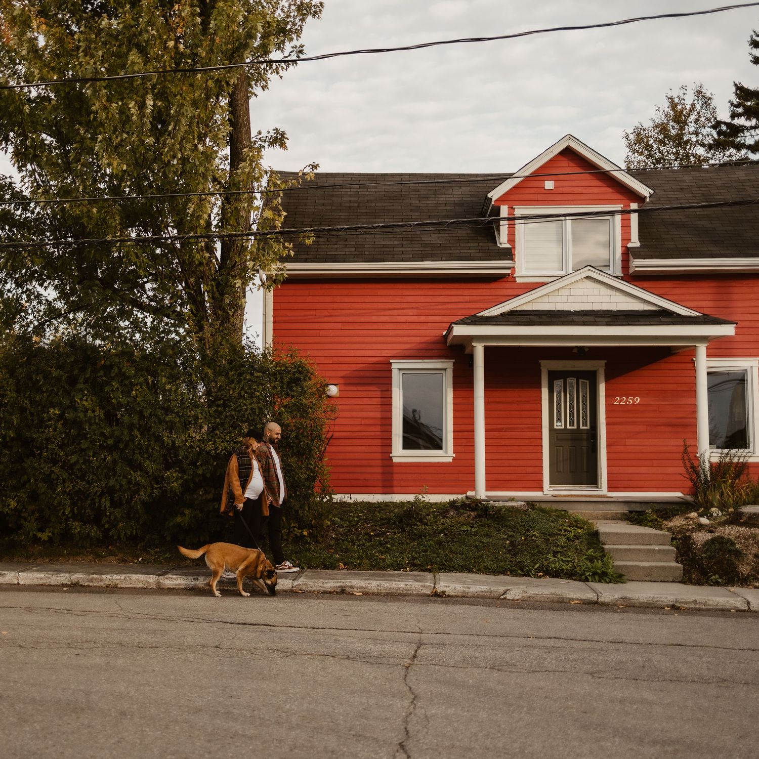 Person walking a dog in front of a charming red two-story house on a quiet residential street.