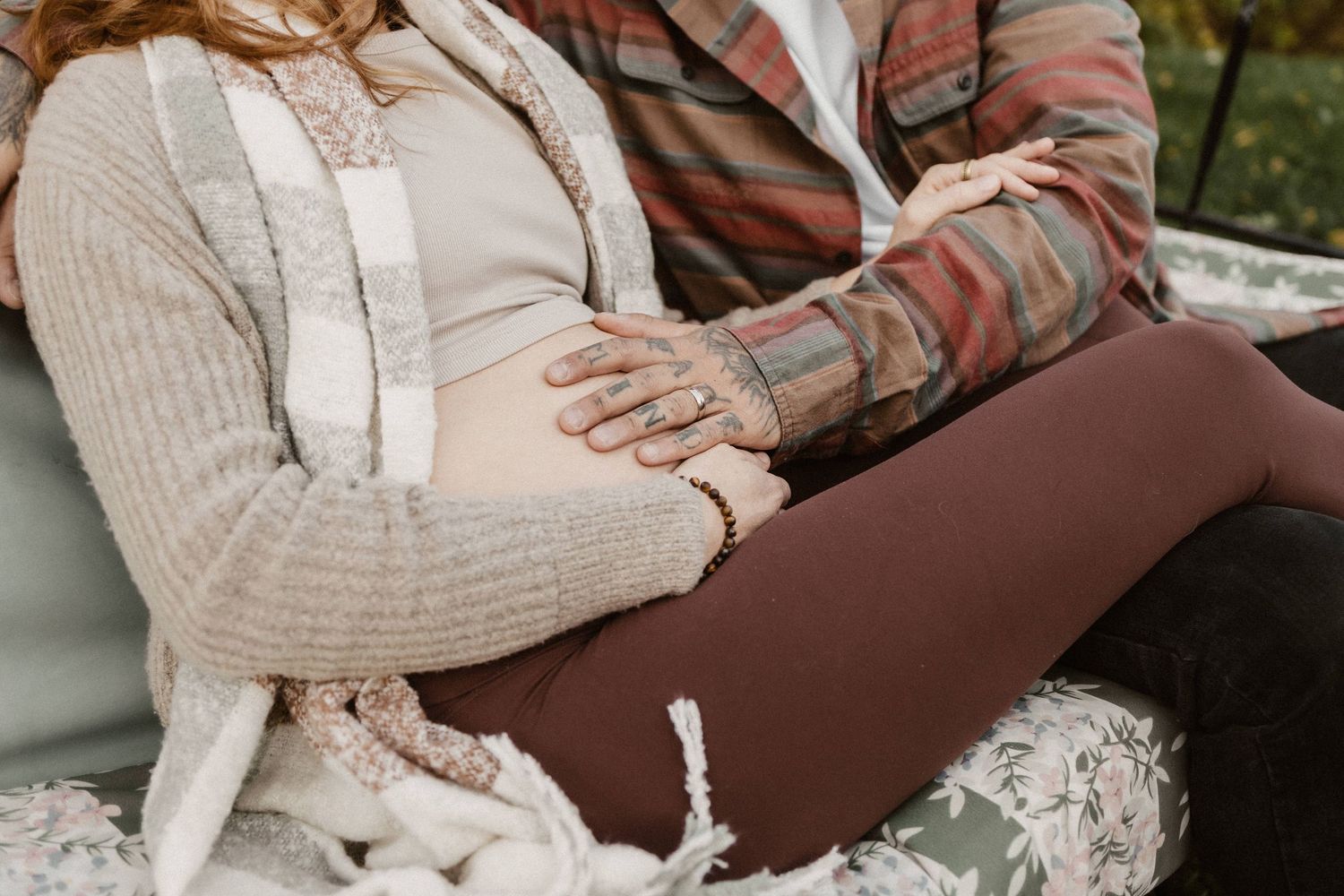 Pregnant woman in cozy cardigan with partner's hand on baby bump during outdoor maternity photo session.