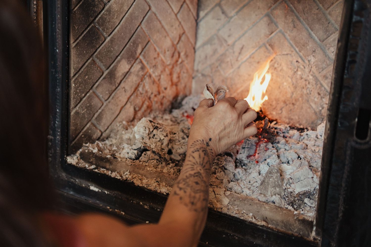 Person with tattooed hand lighting a fire in a brick fireplace with glowing embers and ash.