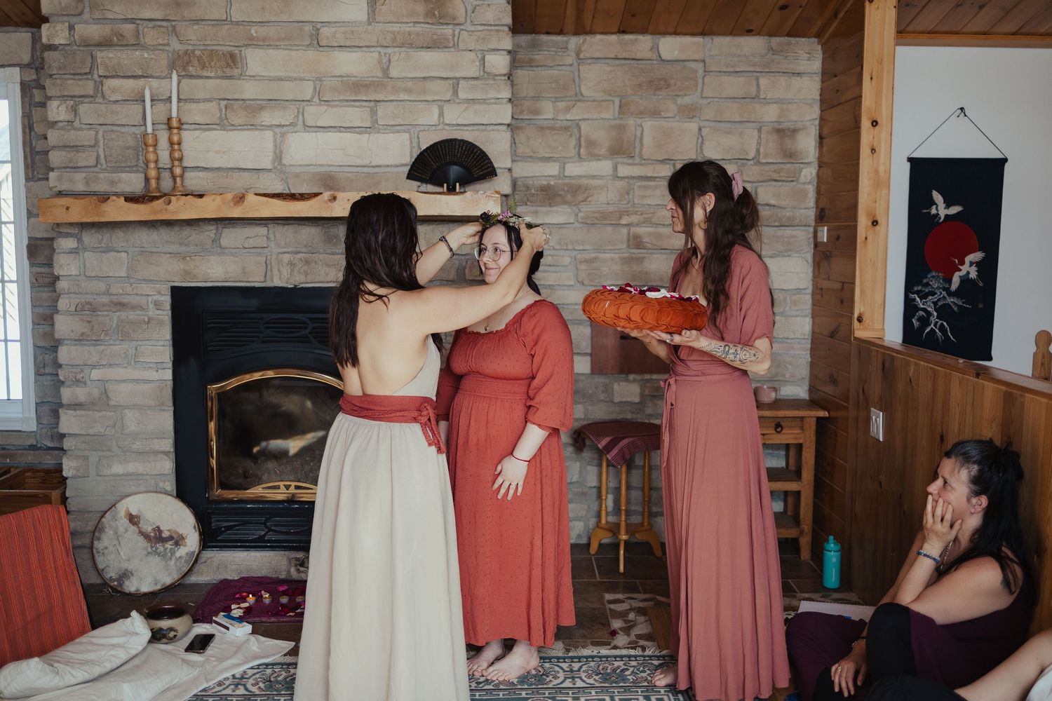 Women in elegant red and white dresses gather near stone fireplace during a ceremonial ritual indoors.
