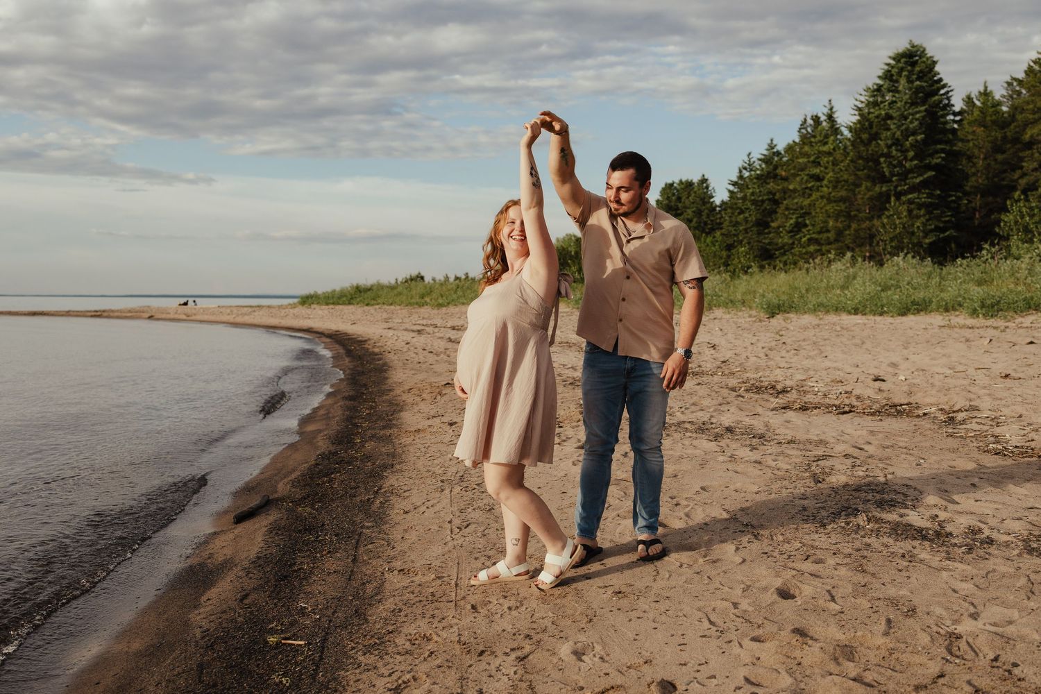 Couple dancing on sandy beach, man spinning pregnant woman near calm water under cloudy sky.