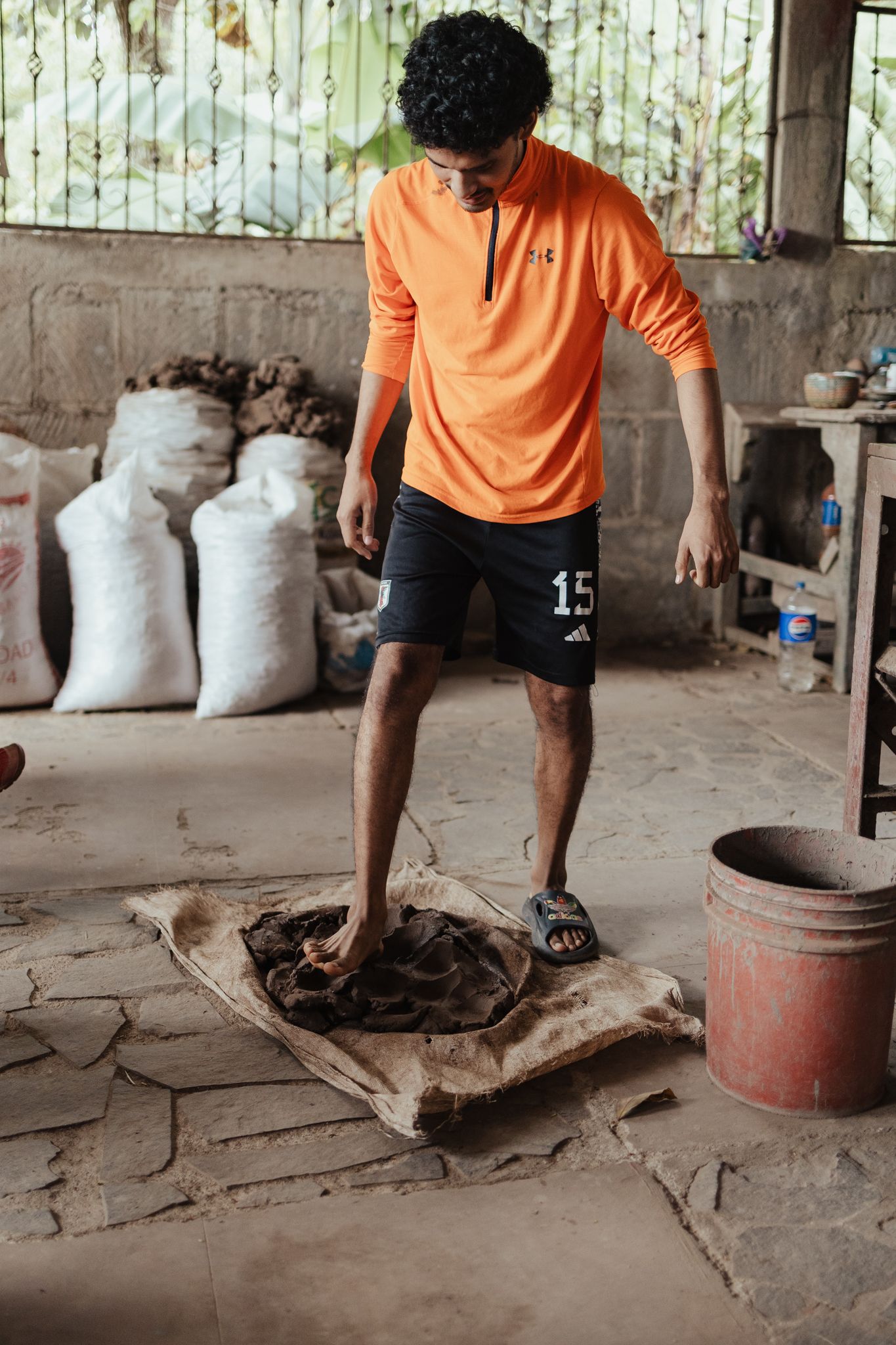 Young man in orange shirt and black shorts standing on burlap sack in rustic workspace with sacks in background.