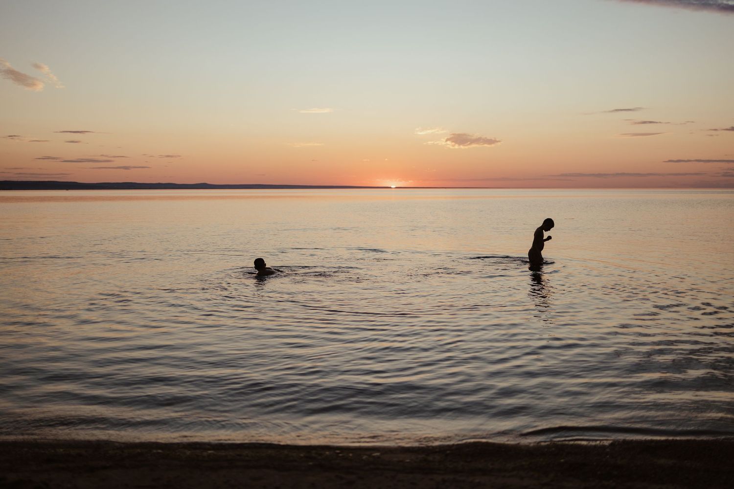 Two silhouettes swimming in calm ocean waters during a stunning orange and pink sunset on a sandy beach.