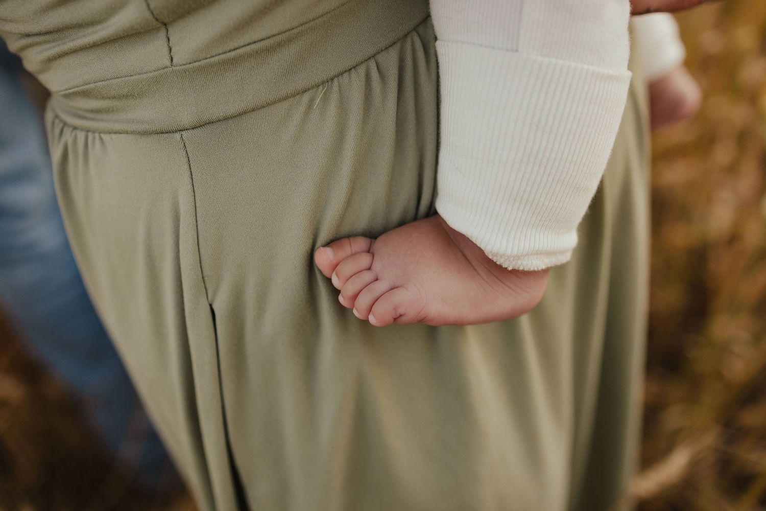 Close-up of a baby's tiny feet resting against a parent's olive green skirt outdoors.