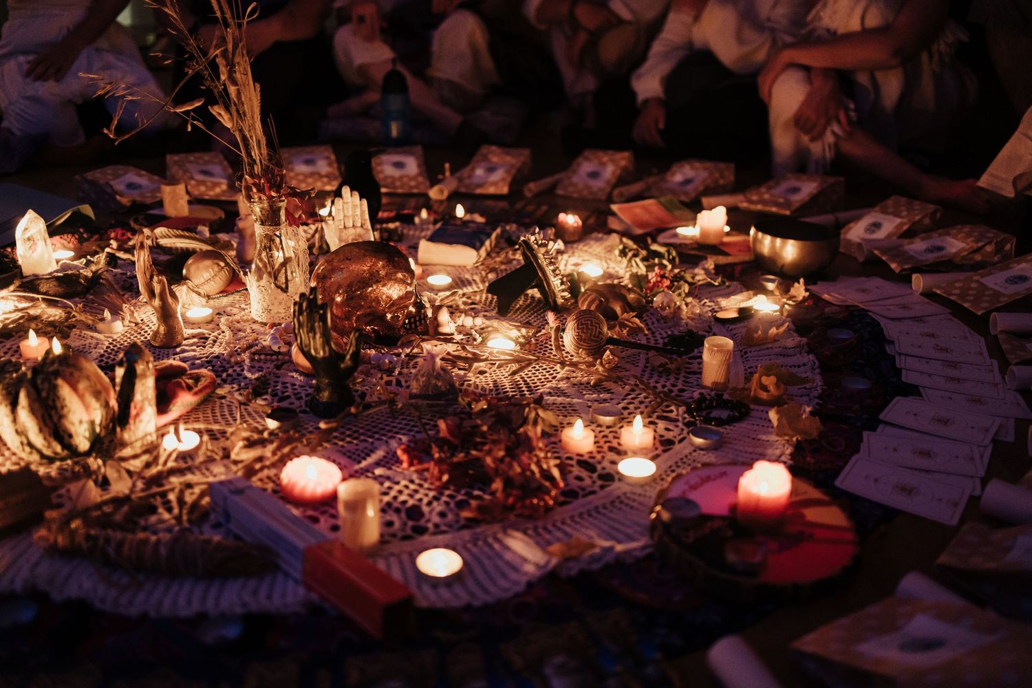 Candlelit ritual altar with flowers, offerings, and decorative items arranged on colorful fabric in a dimly lit ceremonial space.