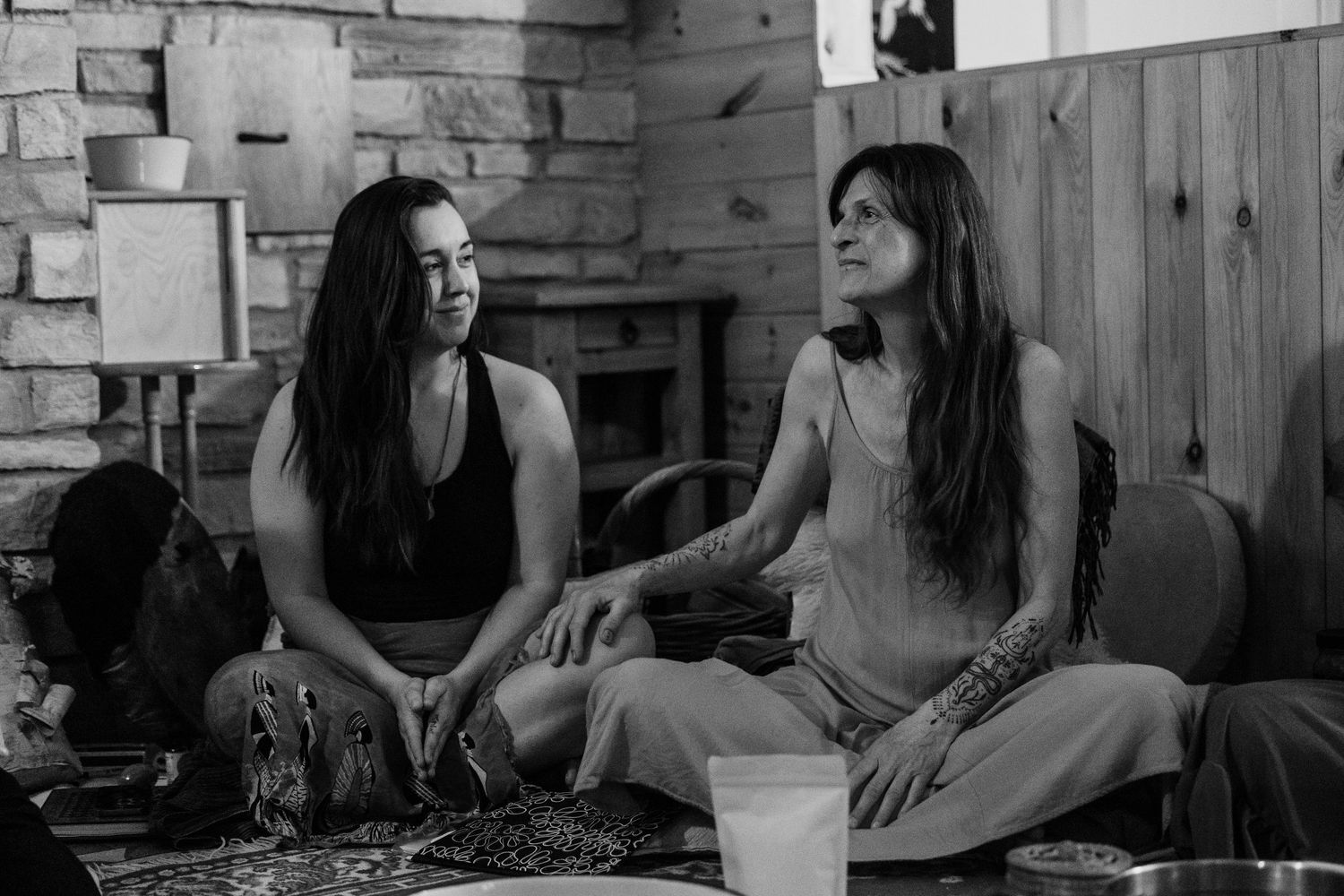 Two women with long dark hair sit cross-legged on the floor, laughing and conversing in a cozy stone-walled room.