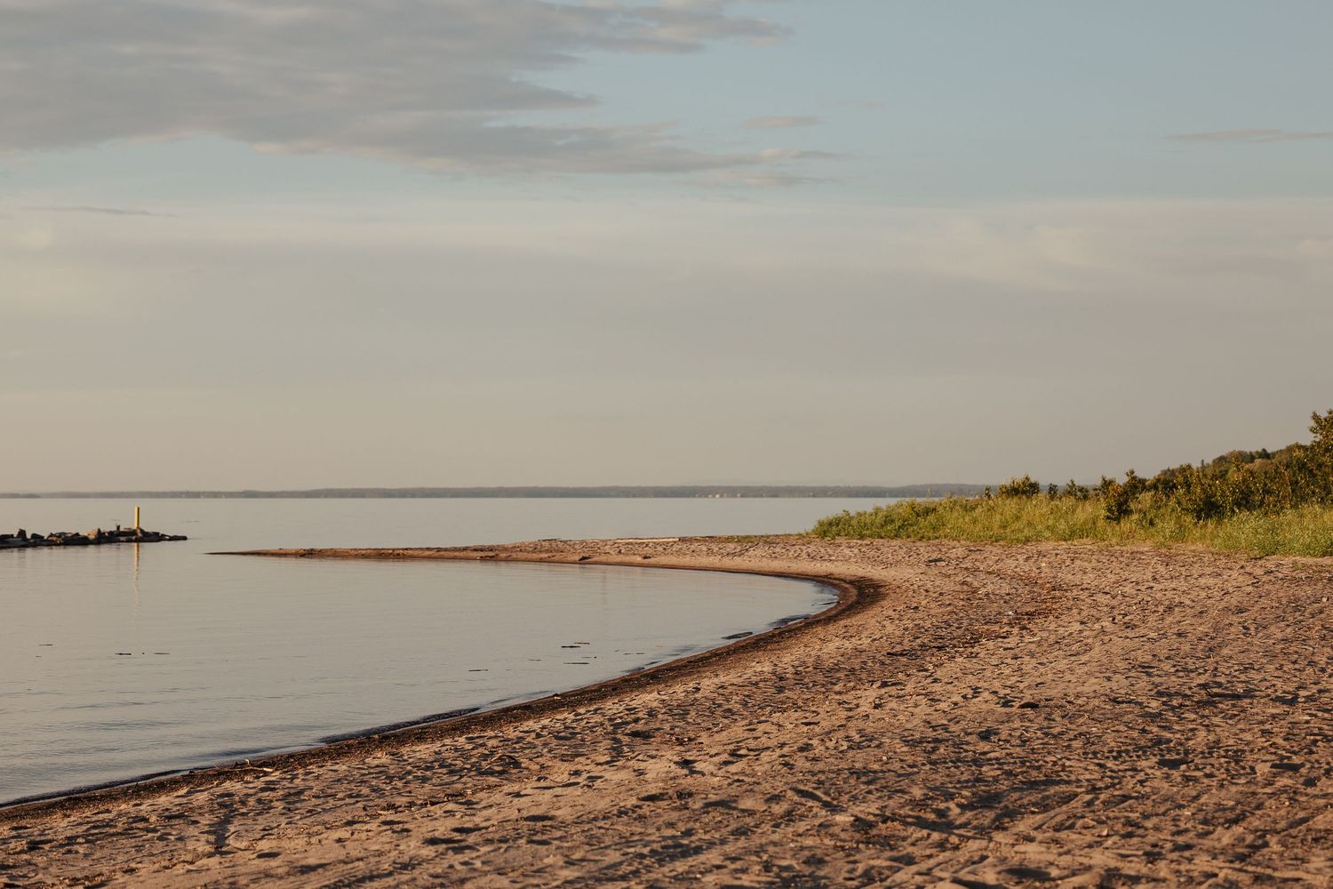 Sandy shoreline curves gently along calm water at golden hour, with lush greenery and soft pastel sky in the background.
