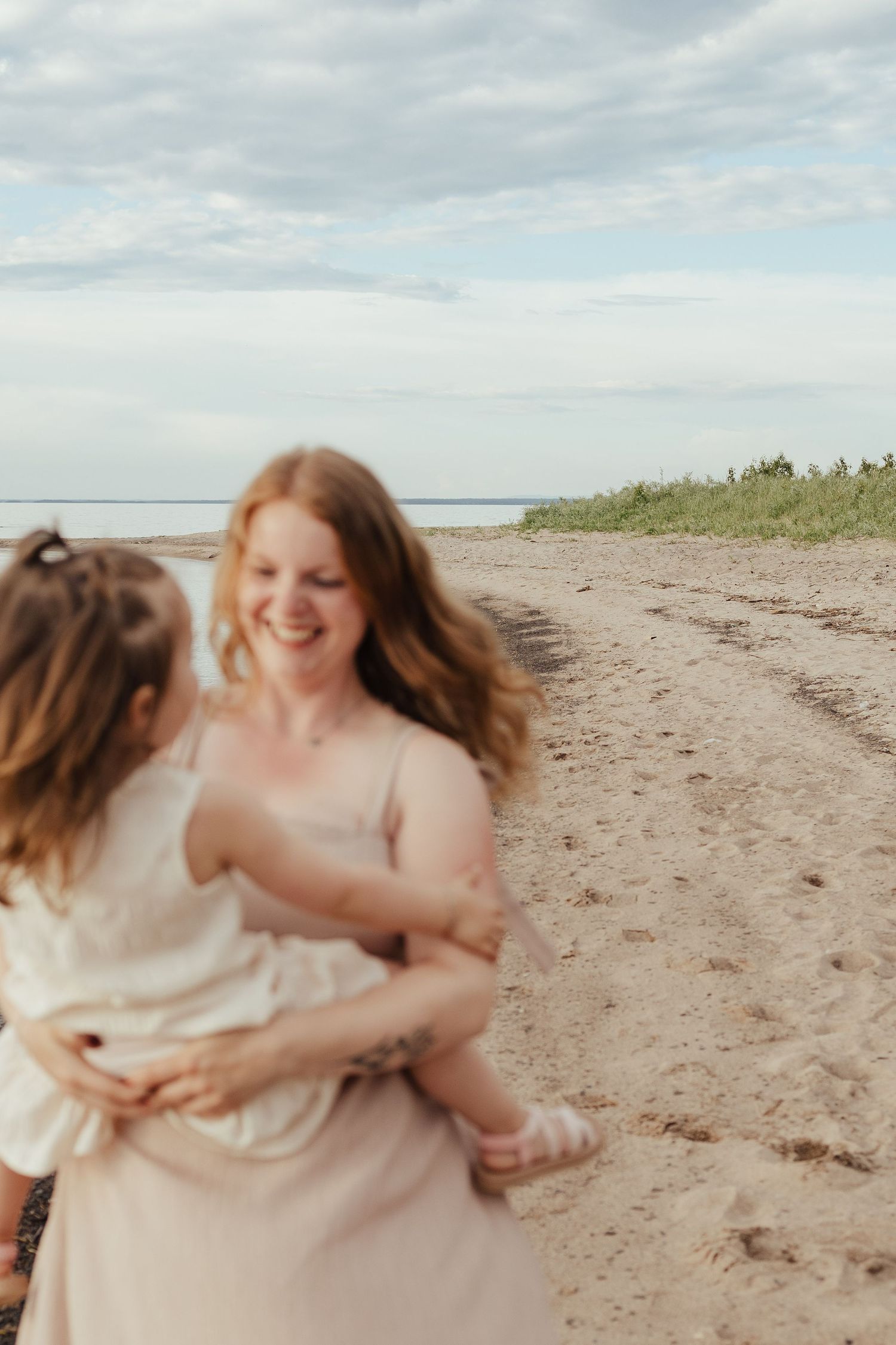 A smiling mother with red hair holds her young daughter on a sandy beach with green dunes and cloudy sky.
