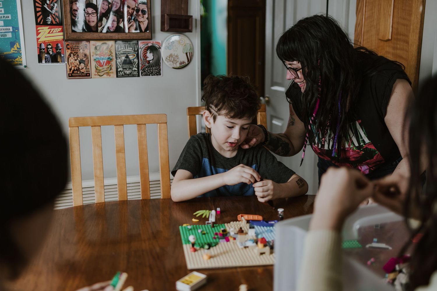 A boy and woman play with LEGO bricks at a wooden dining table, with colorful posters on the wall behind them.