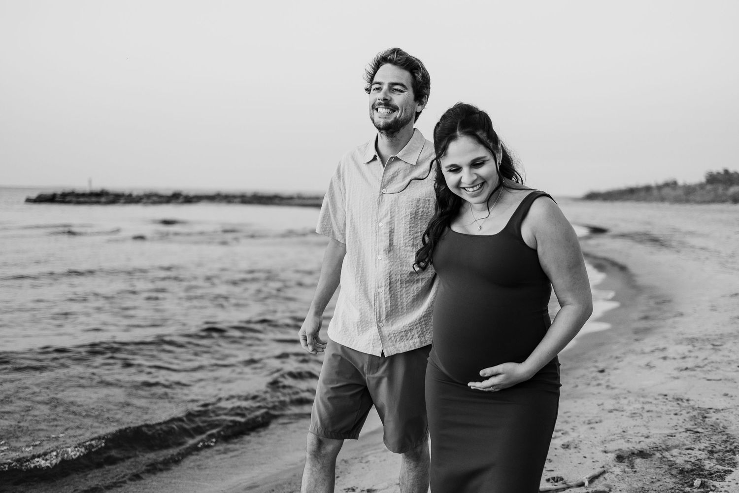 Pregnant couple smiling and walking on a beach at sunset in a black and white maternity photo session.