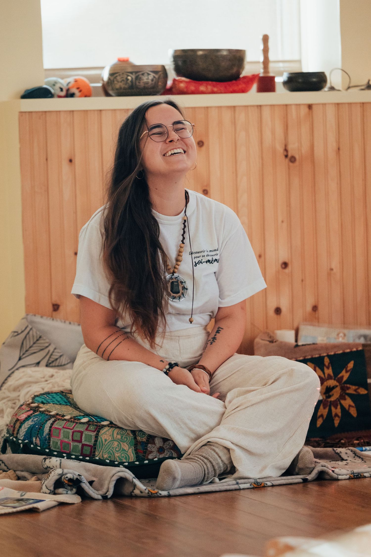 Woman sitting cross-legged on floor cushions, laughing joyfully in a cozy meditation room with singing bowls.