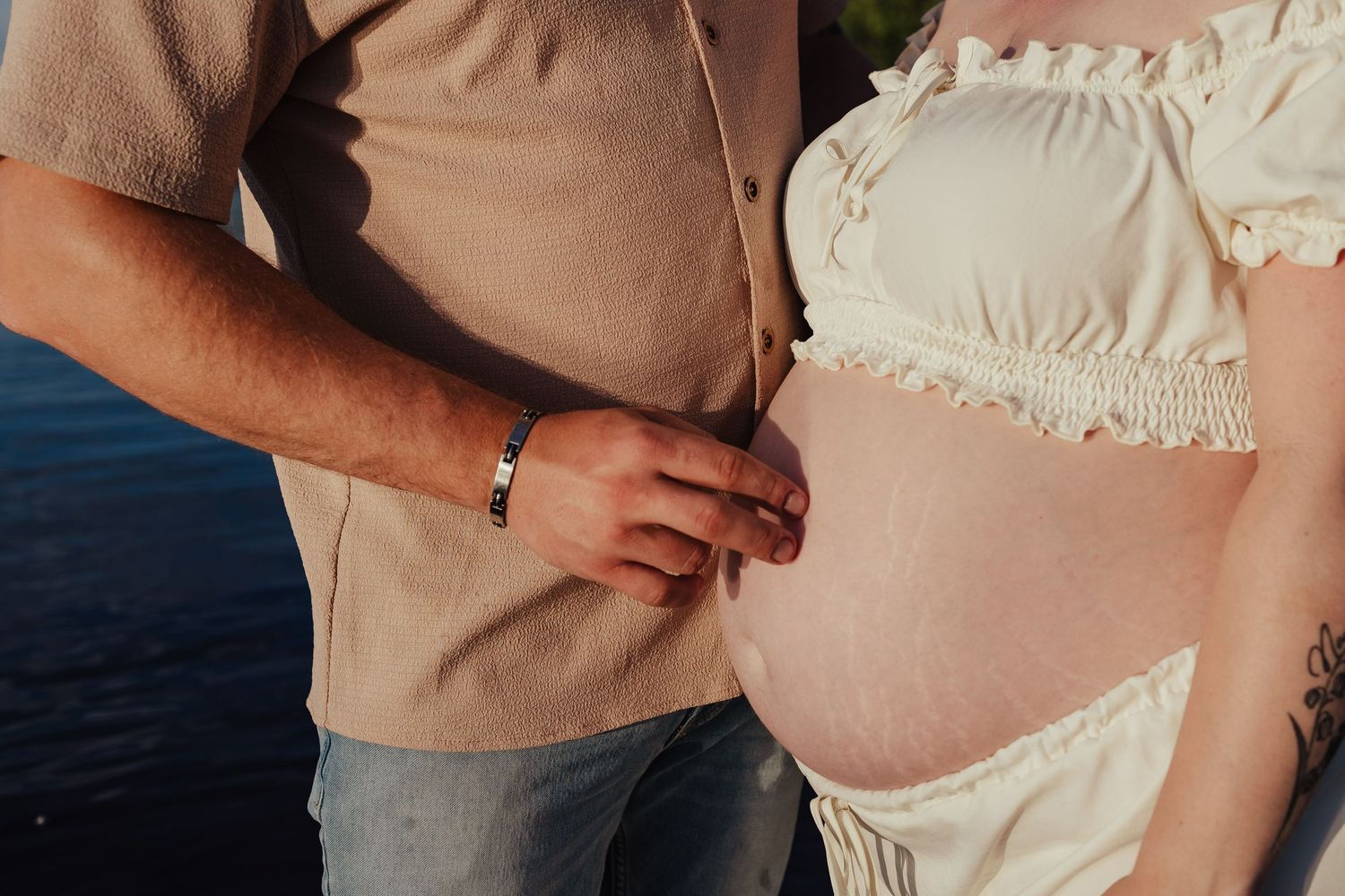 Close-up of man holding hands with pregnant woman showing baby bump in cream ruffled dress outdoors.