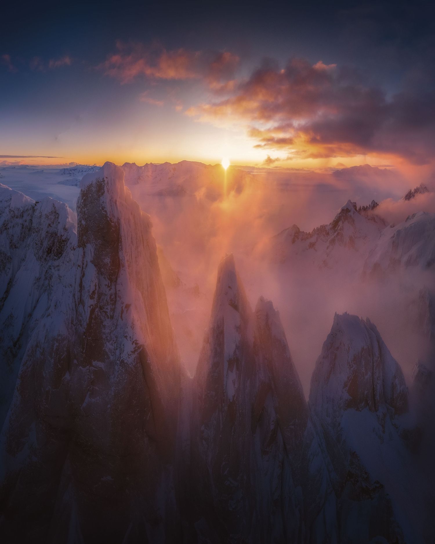 Aerial view of snow-capped mountain peaks bathed in golden sunset light with dramatic clouds and misty valleys below.