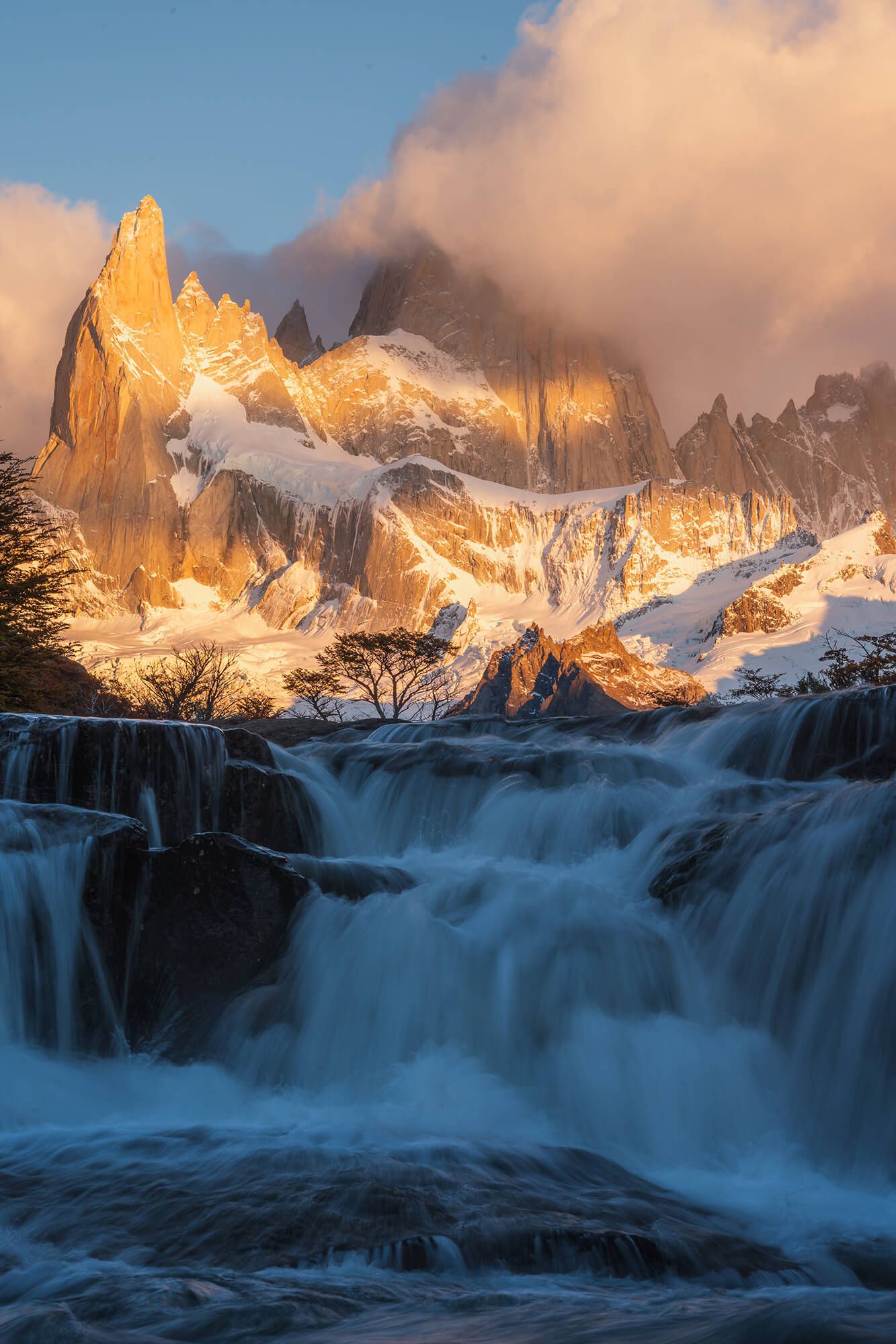 Powerful waterfall cascades before the golden sunlit peaks of Mount Fitz Roy shrouded in clouds, Patagonia.