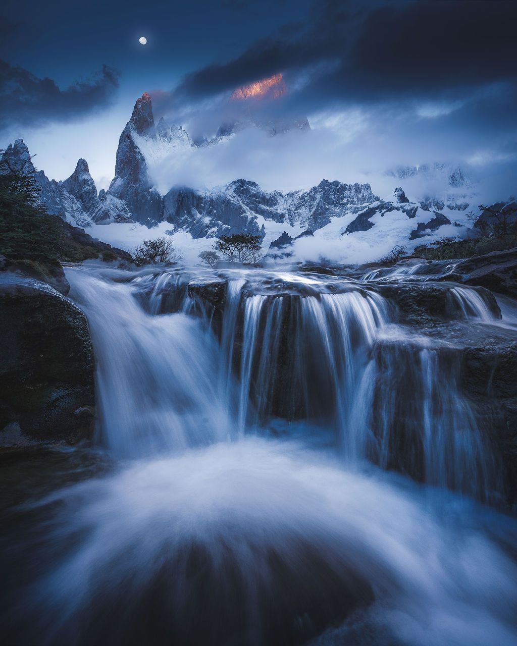 Moonlit waterfall cascades over rocks with misty Fitz Roy mountain peaks in background.