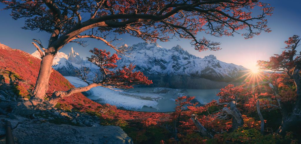 Autumn red trees frame a serene glacial lake with snow-capped mountains glowing in golden sunset light.