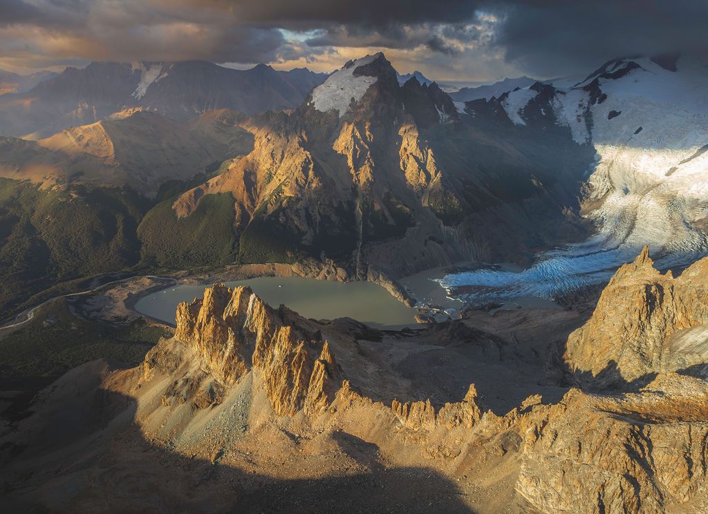 Dramatic aerial view of a glacial lake surrounded by rugged mountain peaks at golden hour.