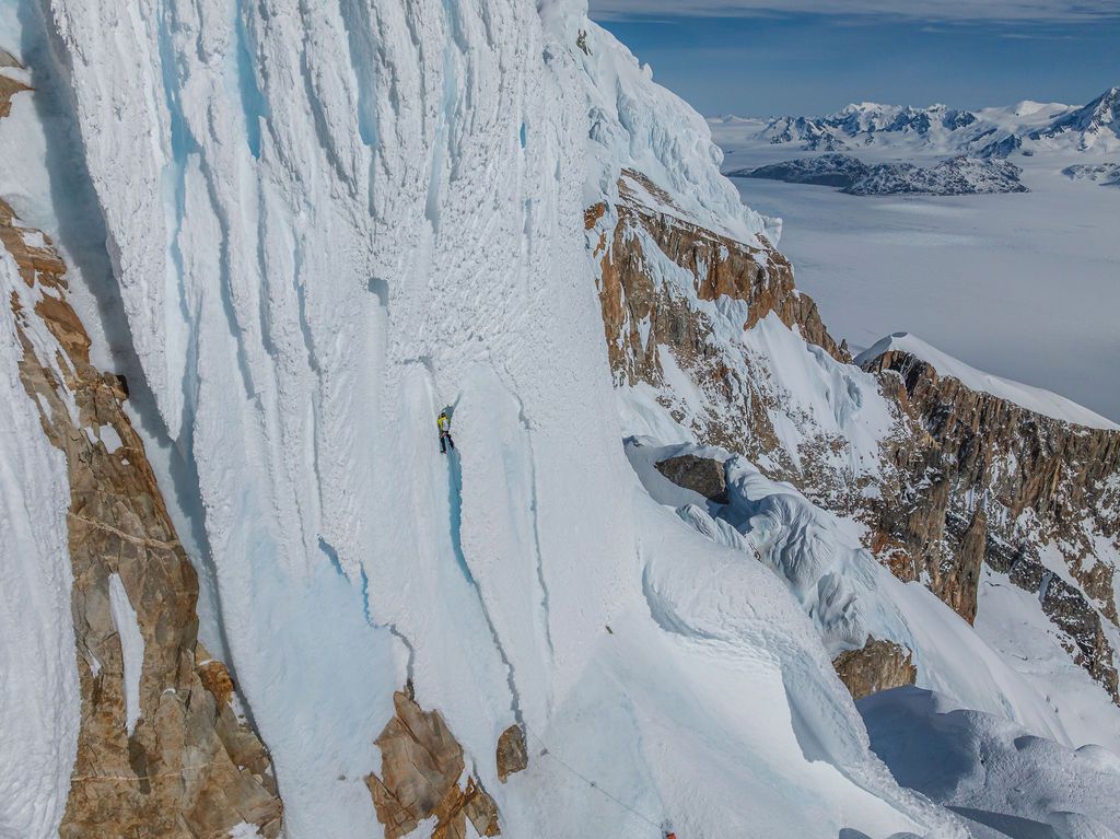 Climber scaling a towering vertical ice wall with frozen landscape and distant mountains below.