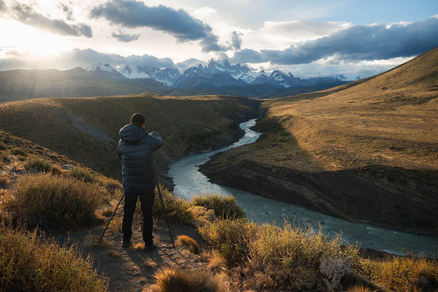 Photographer on tripod captures winding river through golden Patagonian valley with snow-capped peaks at sunset.
