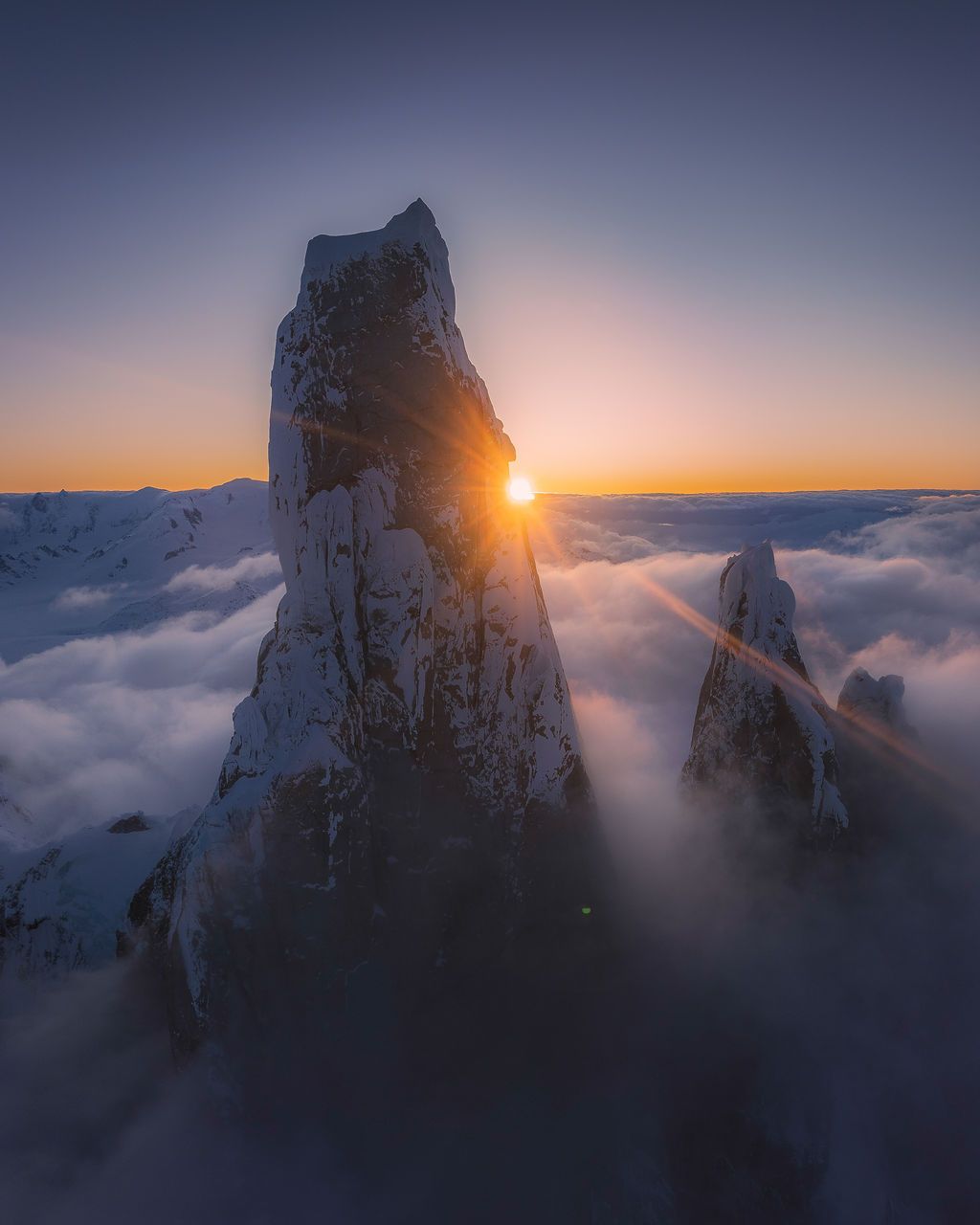 A lone jagged snow-covered rock pinnacle silhouetted against a vivid orange and blue sunset sky.