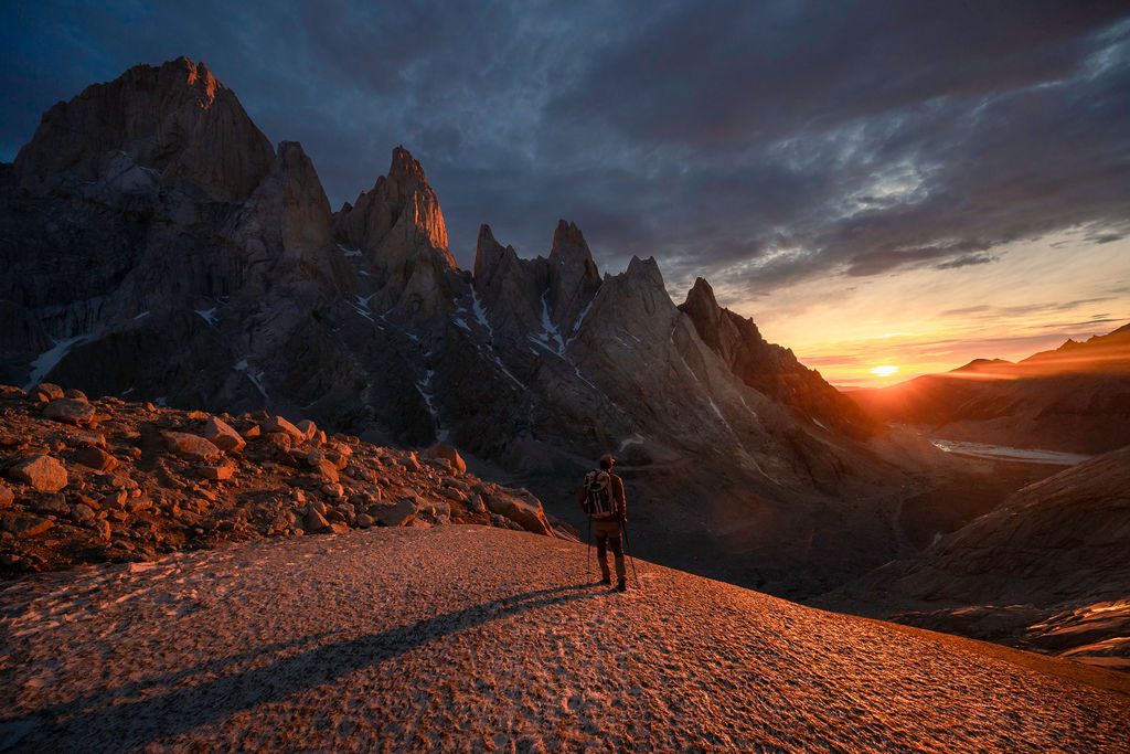 Lone hiker stands before jagged Dolomite peaks bathed in fiery orange sunset light at dusk.