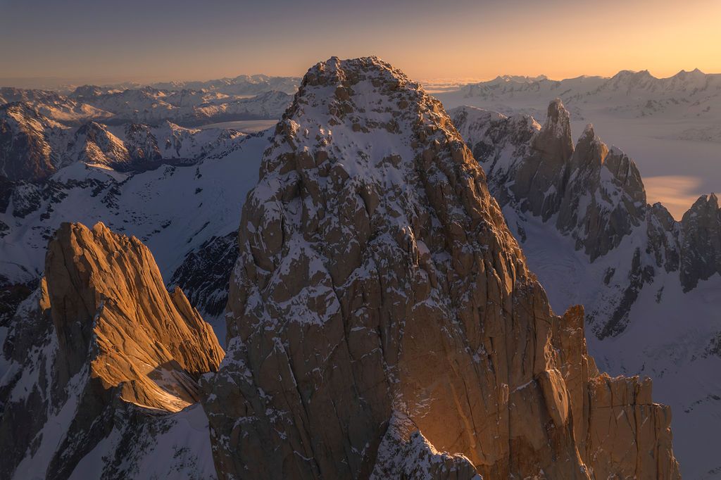 Aerial sunset view of snow-covered rocky mountain peaks glowing in warm golden light.