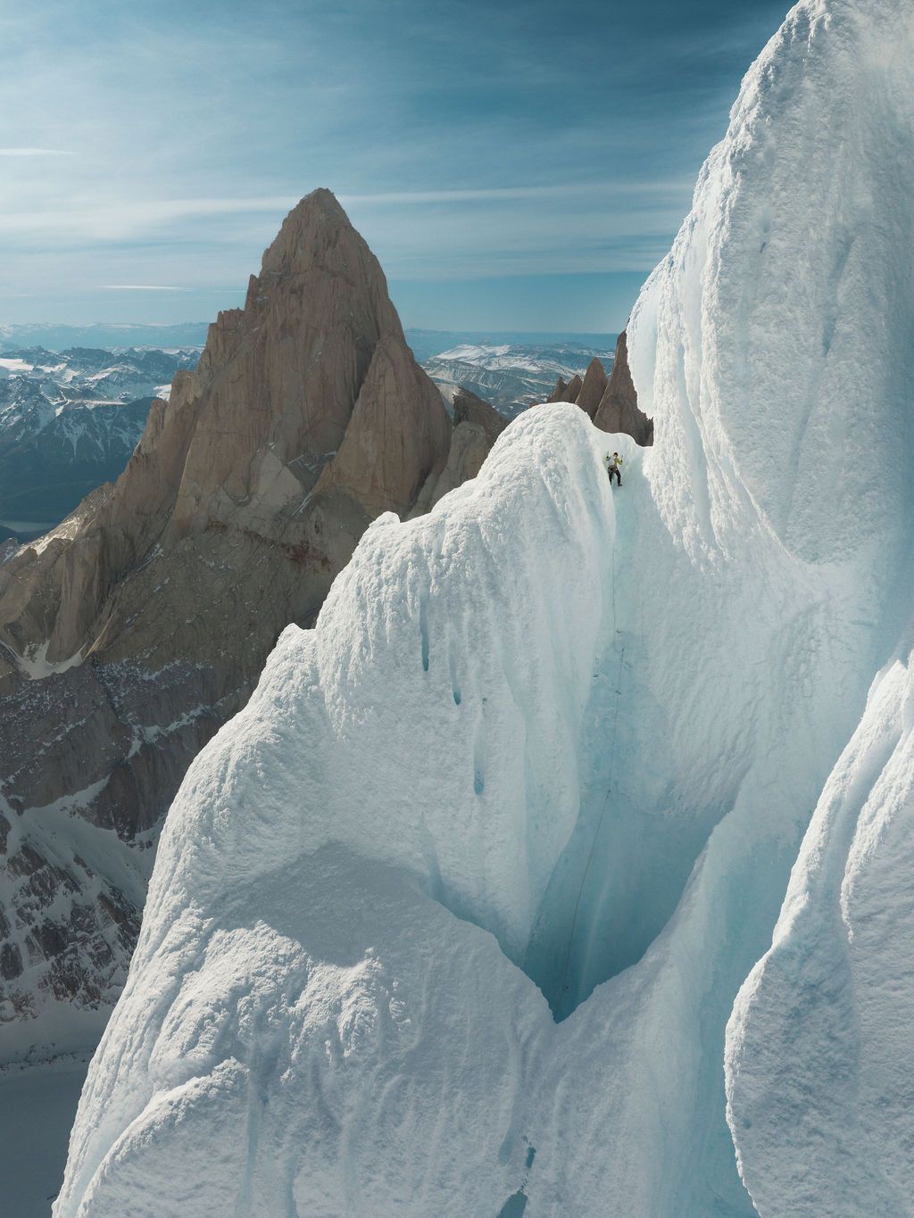 Climber perched on dramatic white ice formation with iconic rocky spire of Fitz Roy in background.