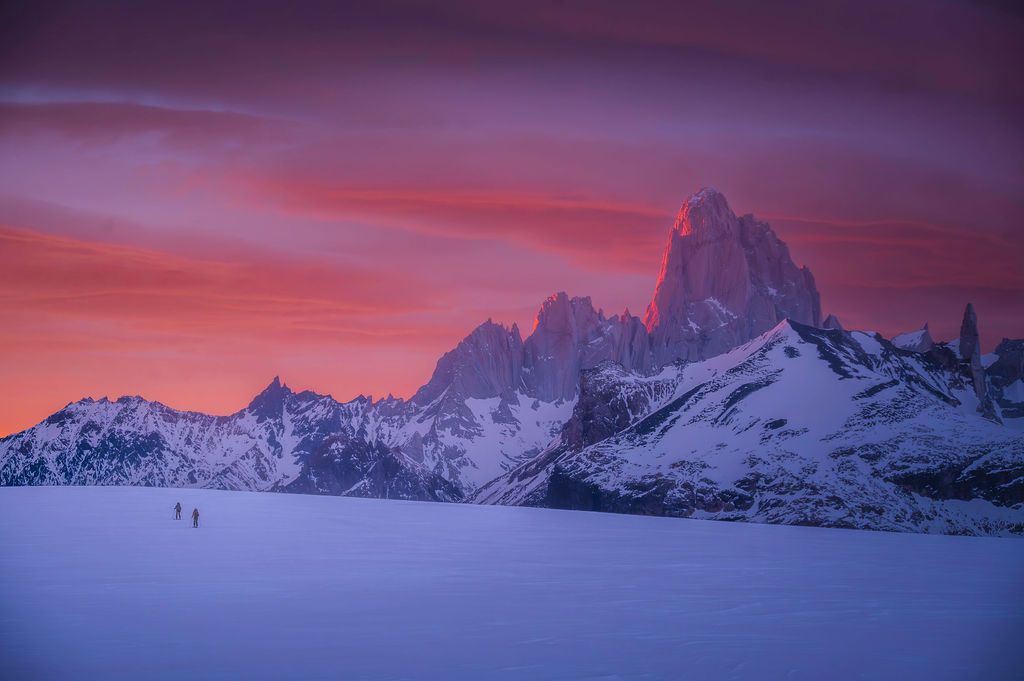 Two tiny figures cross a vast snowy plain beneath a vivid pink sky with Fitz Roy peak in Patagonia.