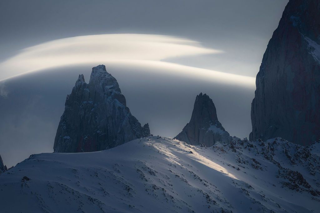 Dramatic lenticular clouds hover over dark jagged mountain peaks in a moody Patagonian winter landscape.