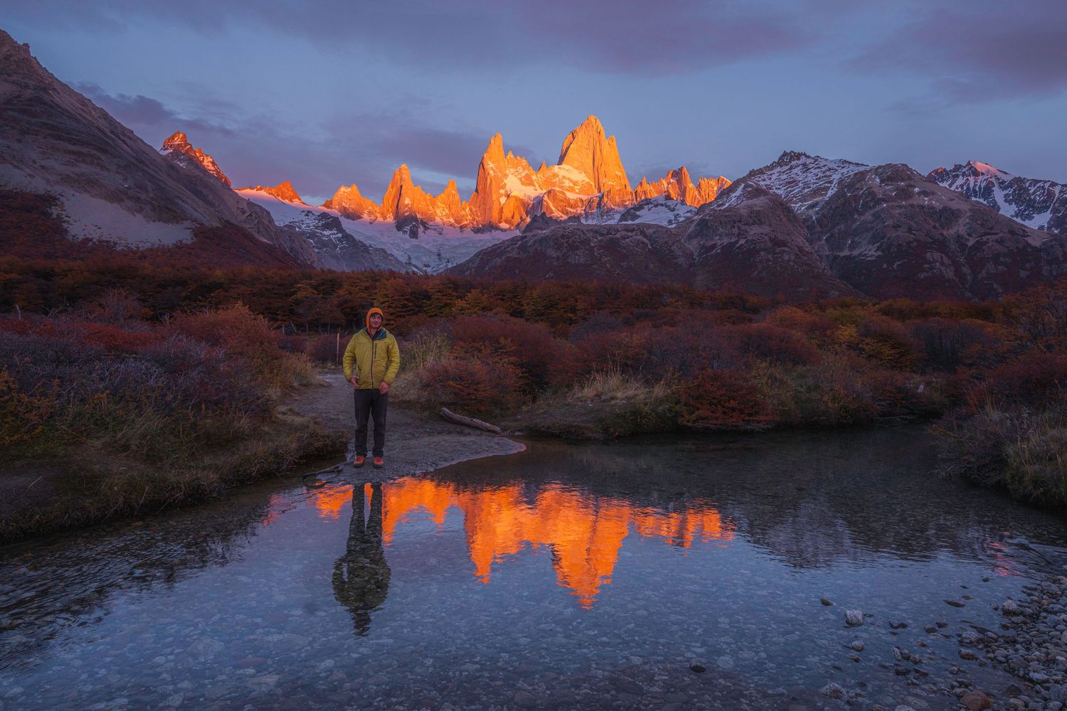 Hiker stands before Mount Fitz Roy at sunrise, reflected in still water, surrounded by autumn foliage in Patagonia.