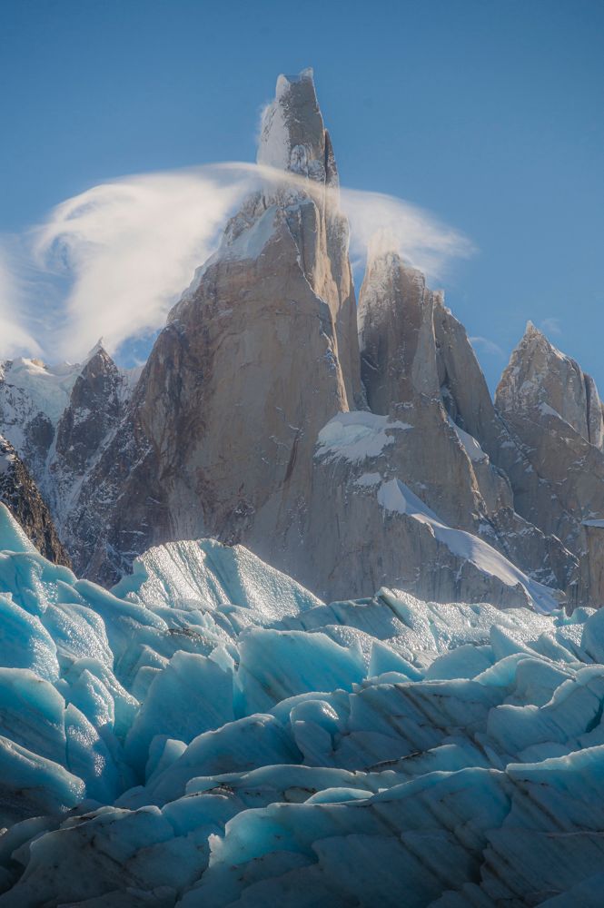 Cerro Torre peak with swirling clouds above a rugged blue glacier in Patagonia, Argentina, under a clear sky.