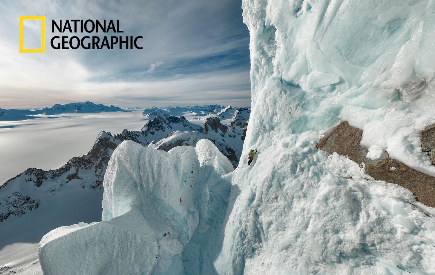 Climber ascending a dramatic ice wall with snowy mountain panorama below.