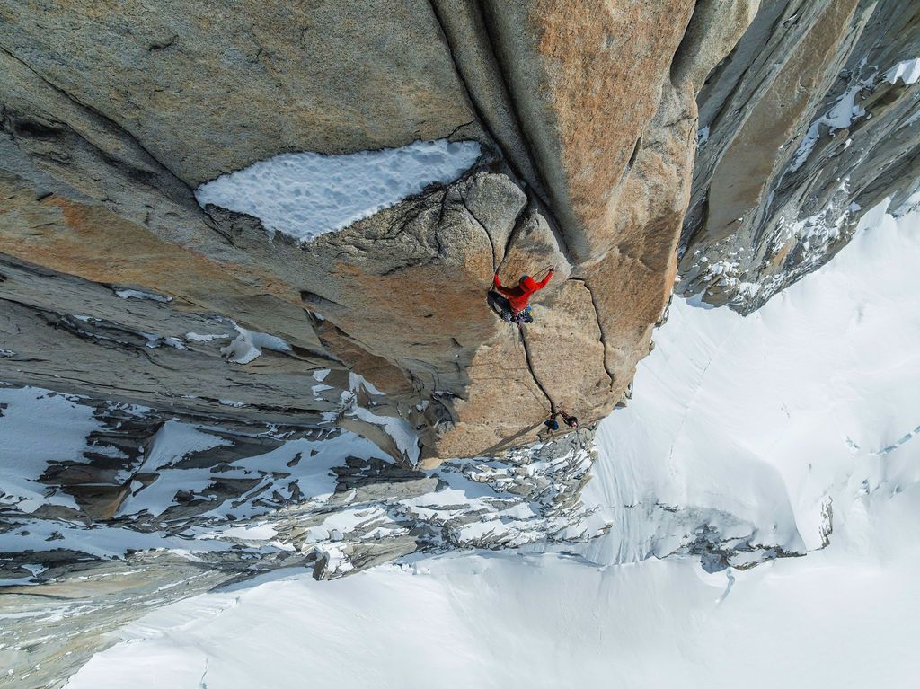 Climber tackles a steep granite crack route with snowy mountain terrain visible below in Patagonia.