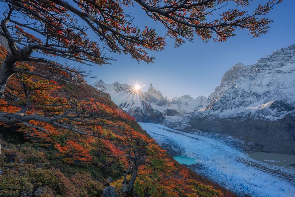 Autumn trees with red foliage frame a sunburst over glacial valley and snowy peaks.